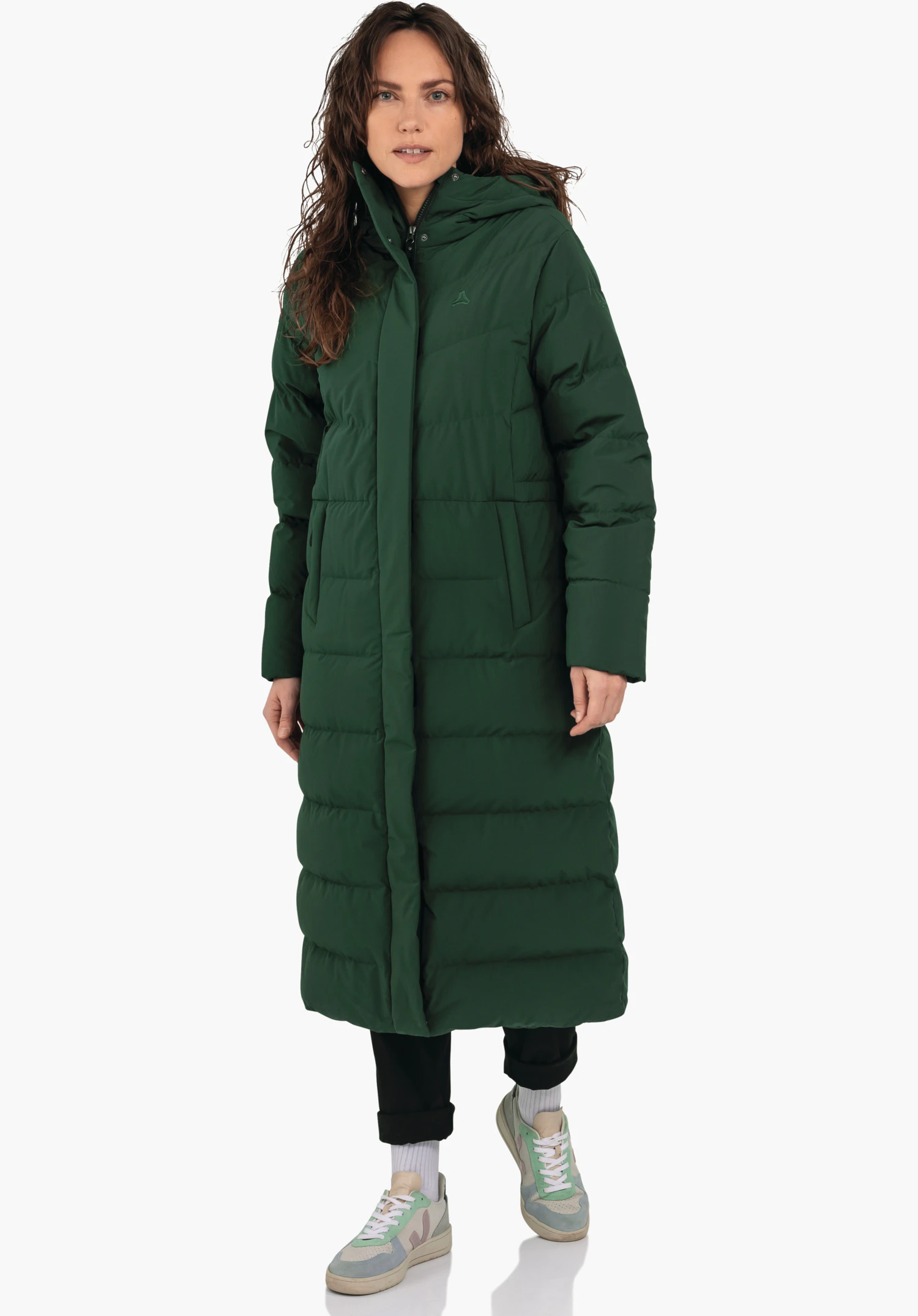 Down Coat Style Vindave WMS dark green long quilted coat worn by a standing woman; hood, side pockets, Schöffel logo on the chest; white background, black trousers, light green and light blue trainers.