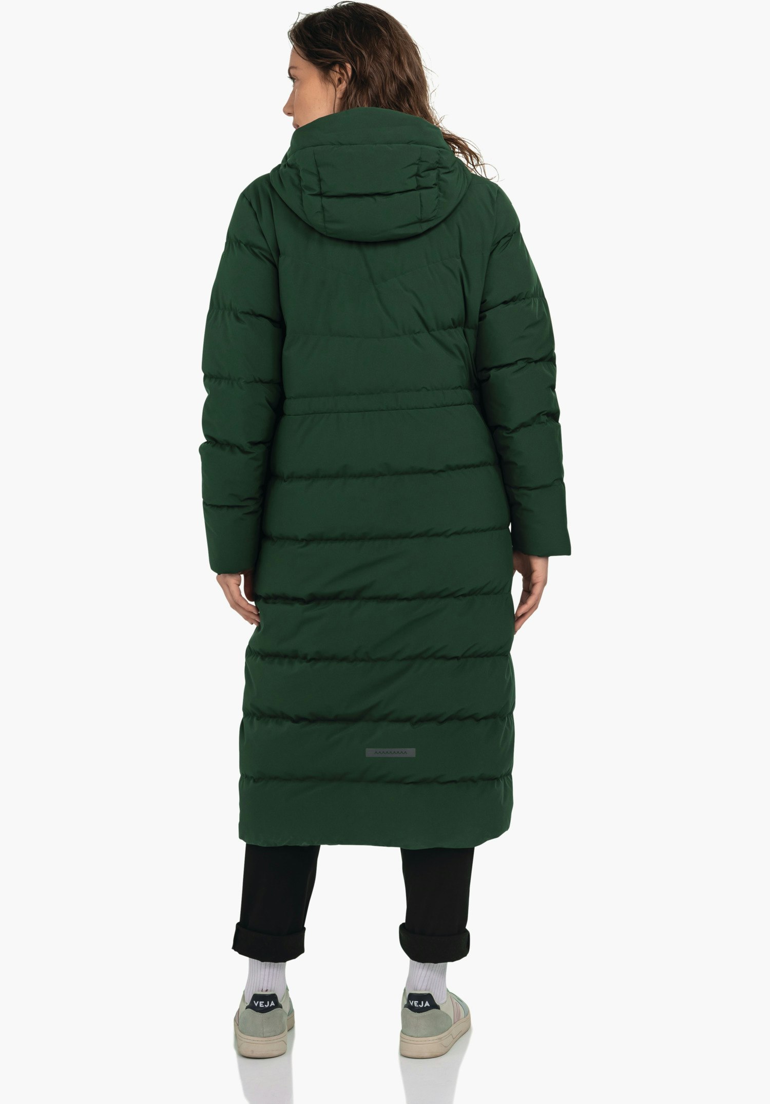 Down Coat Style Vindave WMS dark green long quilted coat worn from the back by a standing person; black trousers, white socks, trainers with “VEJA” text; neutral white studio background.
