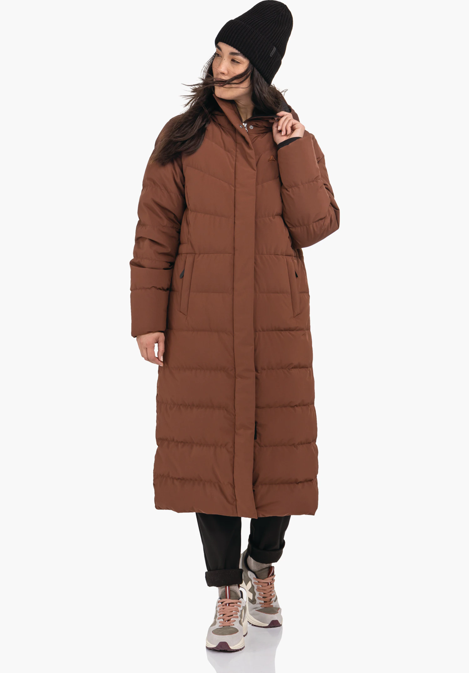 Down Coat Style Vindave WMS lange bruine gewatteerde mantel, gedragen door een persoon die zijwaarts kijkt en de kraag vastpakt; met zwarte muts, zwarte broek en witte sneakers; Schöffel-logo op de borst; witte achtergrond.