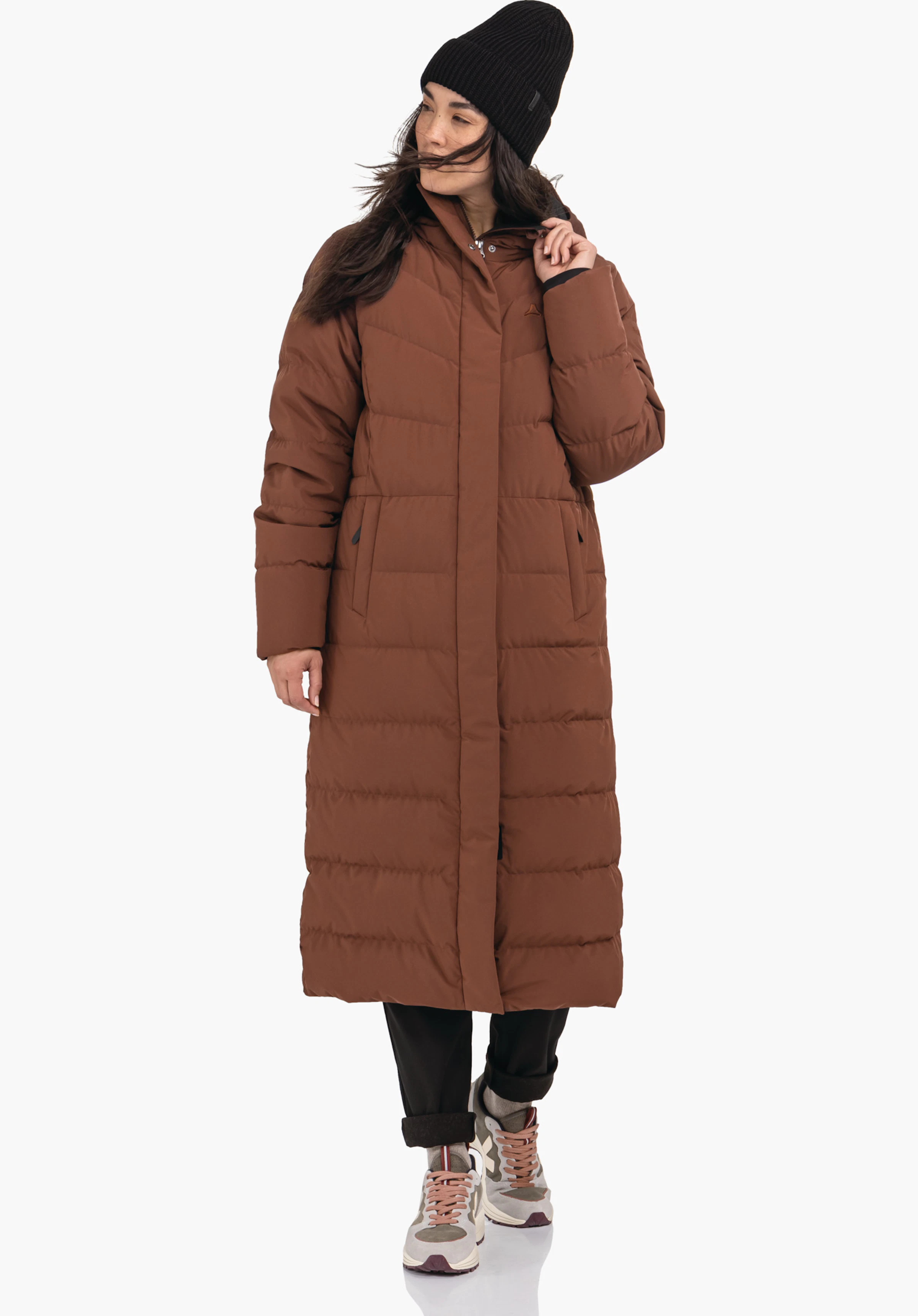 Down Coat Style Vindave WMS lange bruine gewatteerde mantel, gedragen door een persoon die zijwaarts kijkt en de kraag vastpakt; met zwarte muts, zwarte broek en witte sneakers; Schöffel-logo op de borst; witte achtergrond.