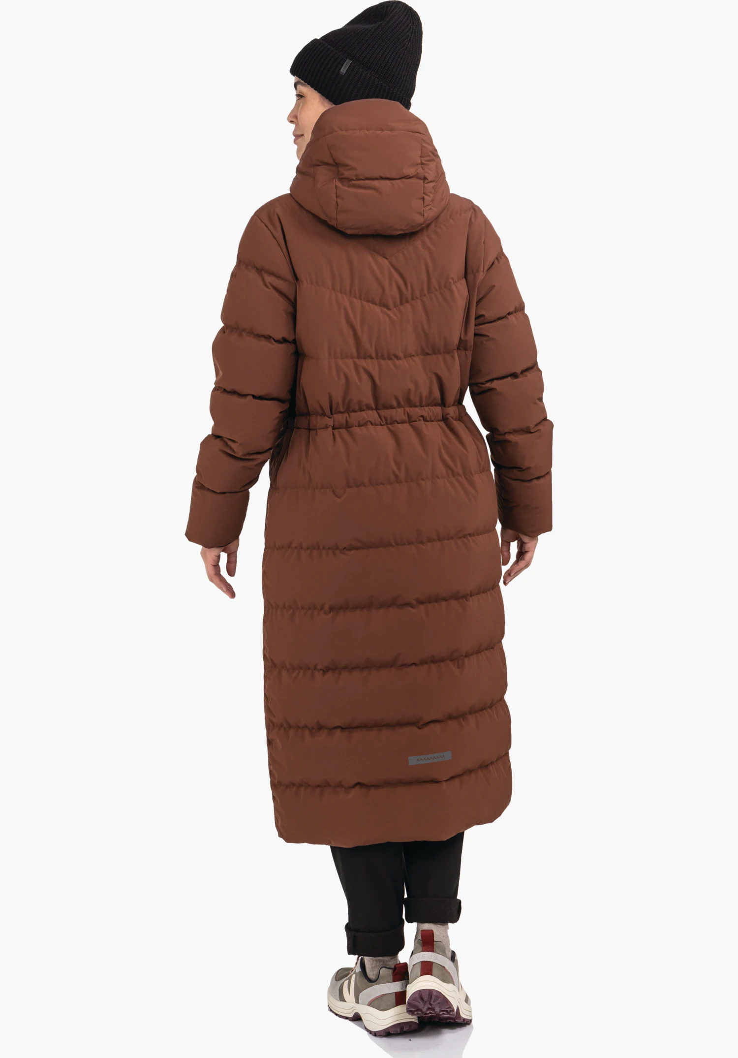 Down Coat Style Vindave WMS lange gewatteerde mantel, gedragen door een persoon die naar links kijkt; witte studioachtergrond, mantel donkerrood, muts zwart, broek zwart, lichte sneakers.