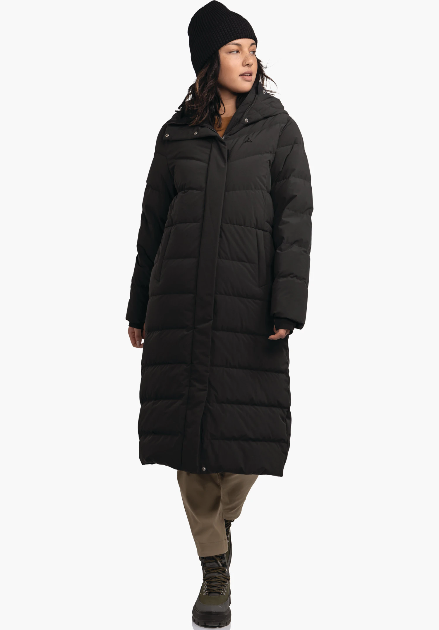 Down Coat Style Vindave WMS long black quilted coat with Schöffel logo; woman wears it while walking; black beanie, brown trousers, green boots; light neutral background.