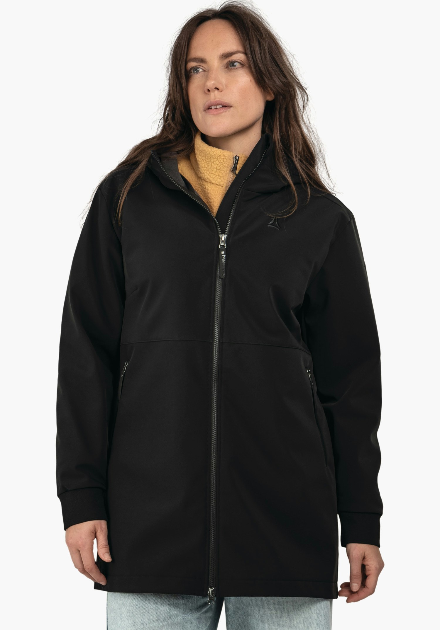 Softshell Jk Style Suavio WMS parka noir longueur genou avec fermeture éclair fermée, portée sur un pull polaire jaune clair; logo Schöffel sur la poitrine; modèle regardant légèrement sur le côté devant un fond blanc.