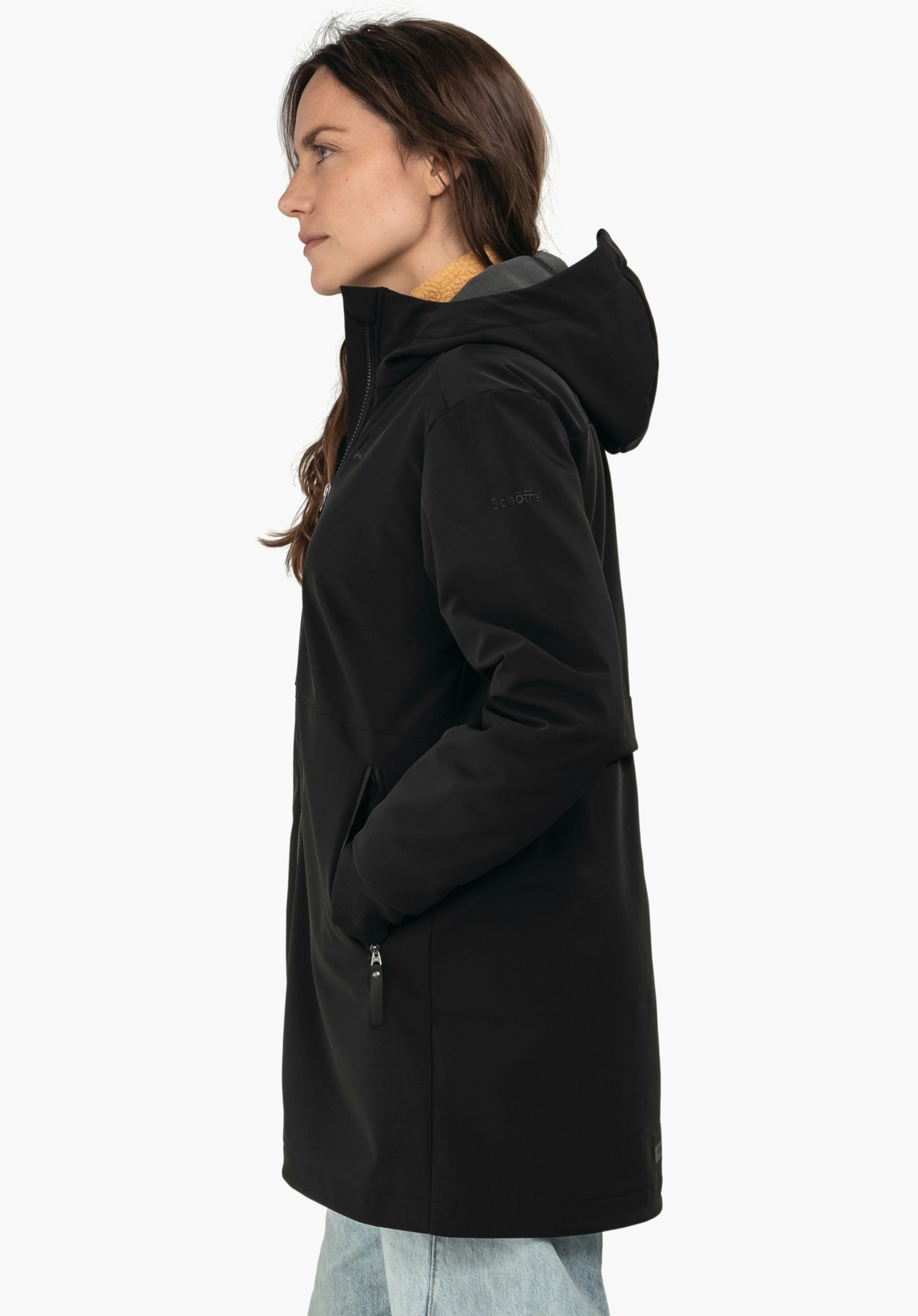 Softshell Jk Style Suavio WMS manteau à capuche noir, femme debout en profil avec les mains dans les poches, col jaune clair visible, porte un jean bleu clair, fond blanc neutre. Texte sur la manche: "Schöffel".