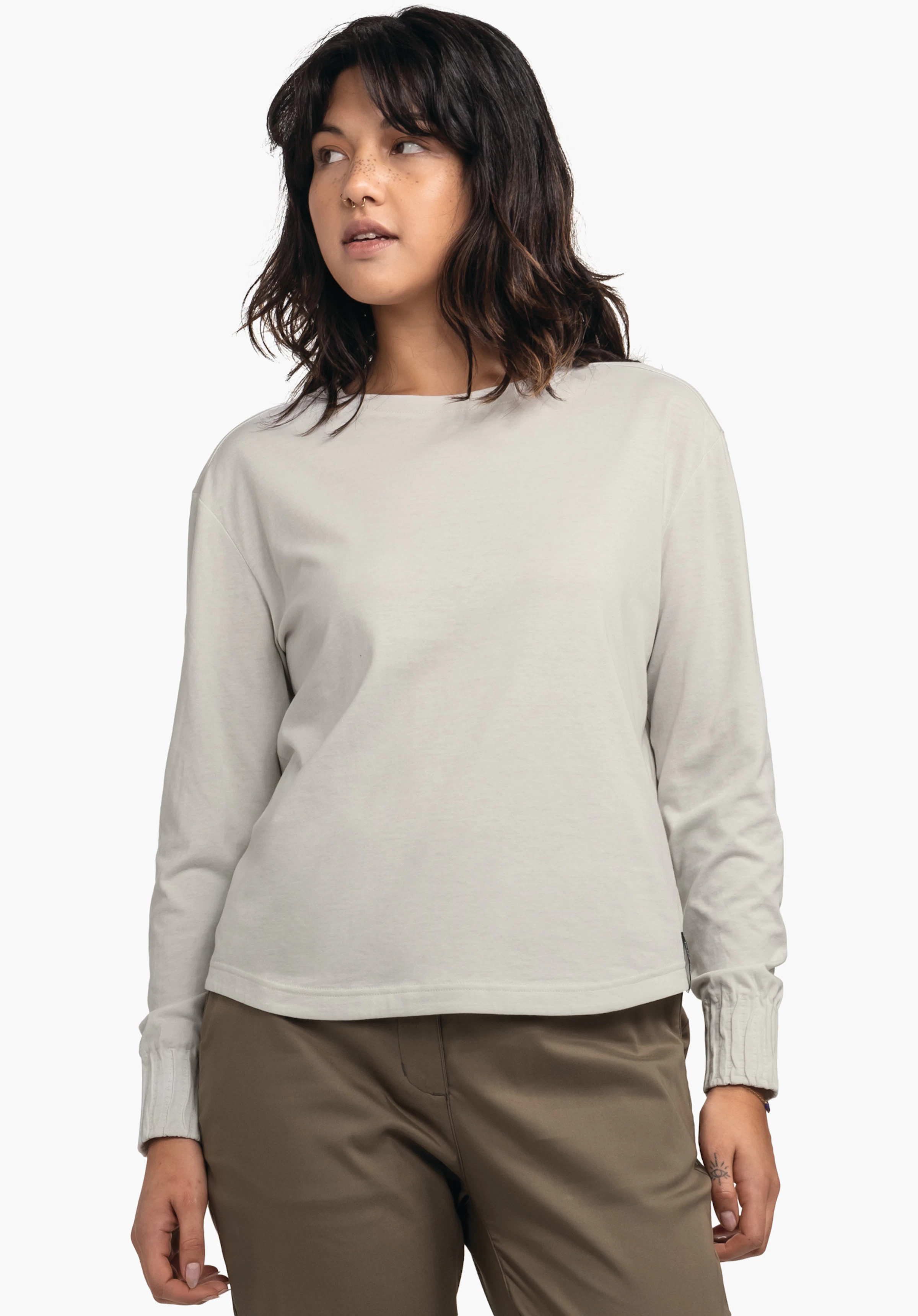 Longsleeve Style Collada WMS Frau trägt hellgraues Langarmshirt, steht leicht seitlich und blickt nach links; Hände locker an den Seiten, trägt dunkelbraune Hose und dunkelbraune Haare; neutraler weißer Hintergrund.