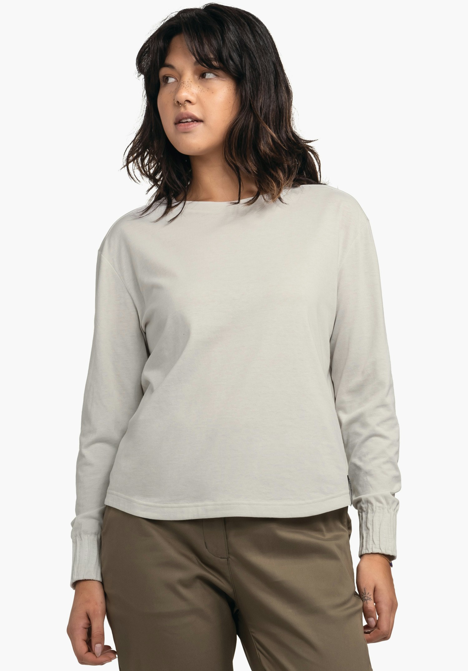 Longsleeve Style Collada WMS woman wearing a light grey long-sleeve top, standing slightly sideways and looking left; arms relaxed, dark brown trousers, dark brown hair; neutral white background.