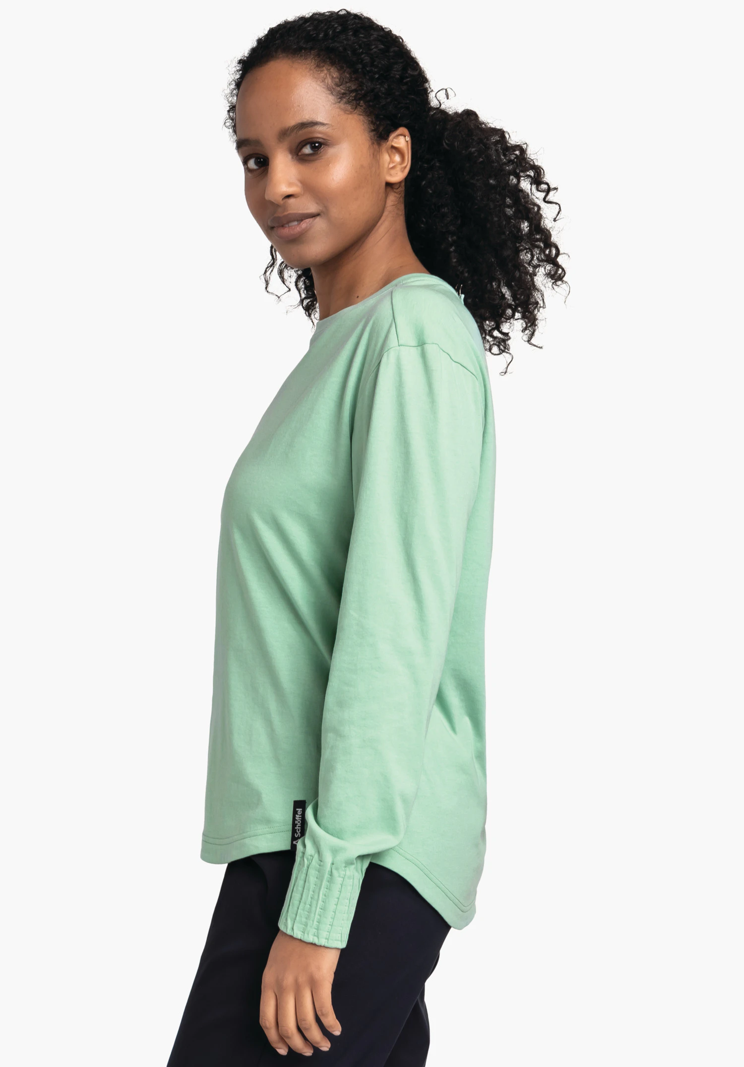 Longsleeve Style Collada WMS t-shirt manches longues vert clair; personne debout de profil regardant la caméra; cheveux bouclés attachés; pantalon bleu foncé; fond blanc neutre.
