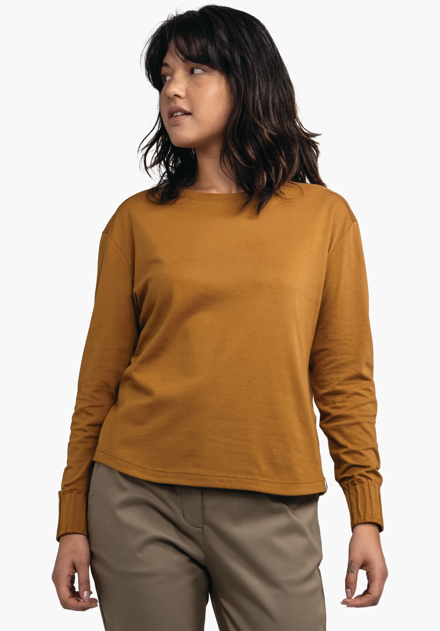 Longsleeve Style Collada WMS femme portant un haut manches longues orange foncé, légèrement de profil et regardant vers la gauche; pantalon gris clair, cheveux noirs; fond blanc.