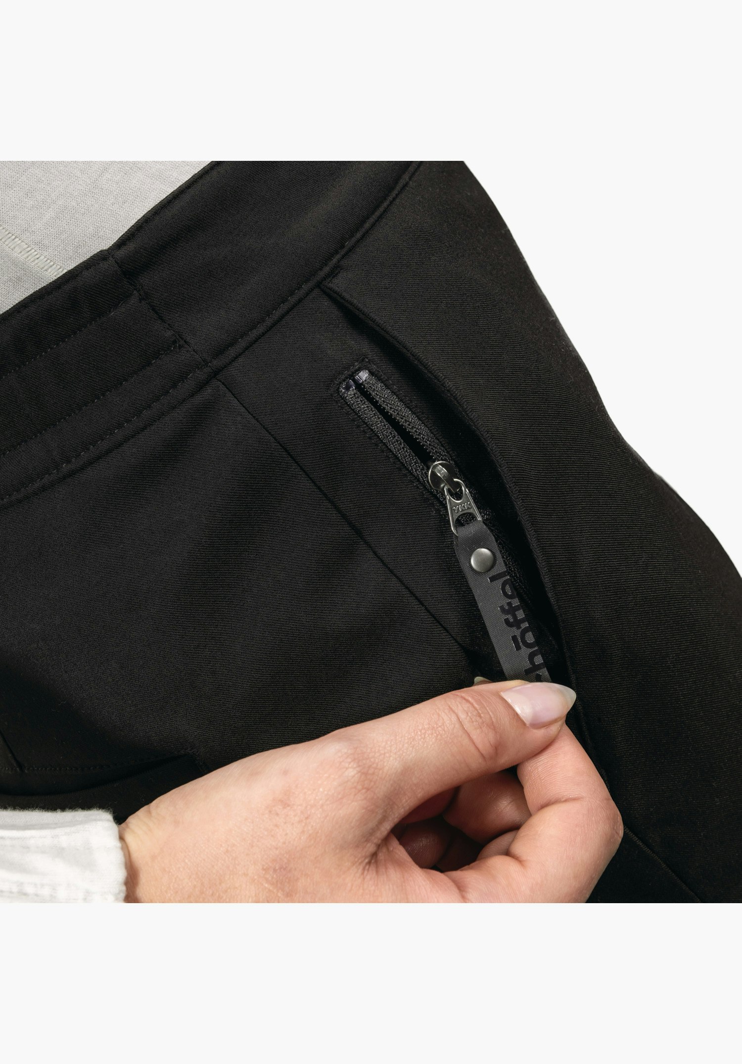 Pants Style Zourga WMS montre la poche zippée latérale en tissu noir, avec une main tenant la tirette allongée portant l'inscription Schöffel, gros plan sur fond blanc.