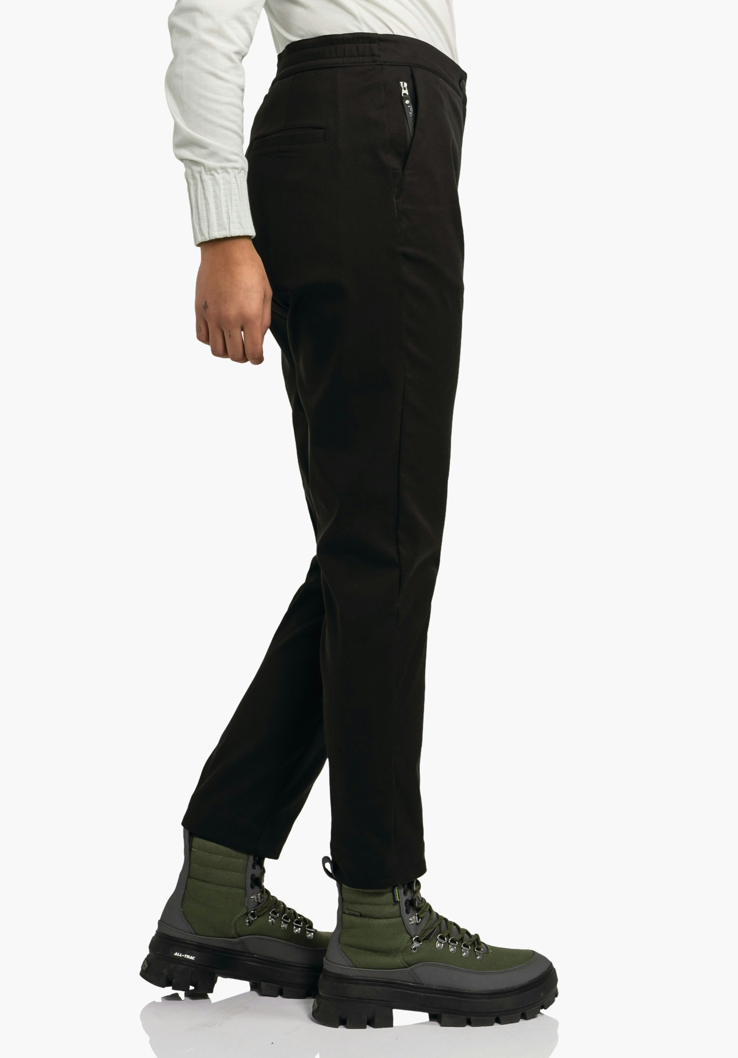 Pants Style Zourga WMS pantalon noir porté de profil, jambe légèrement croisée; main détendue sur le côté; contexte: fond blanc; haut blanc clair; bottes vert foncé à semelles noires.