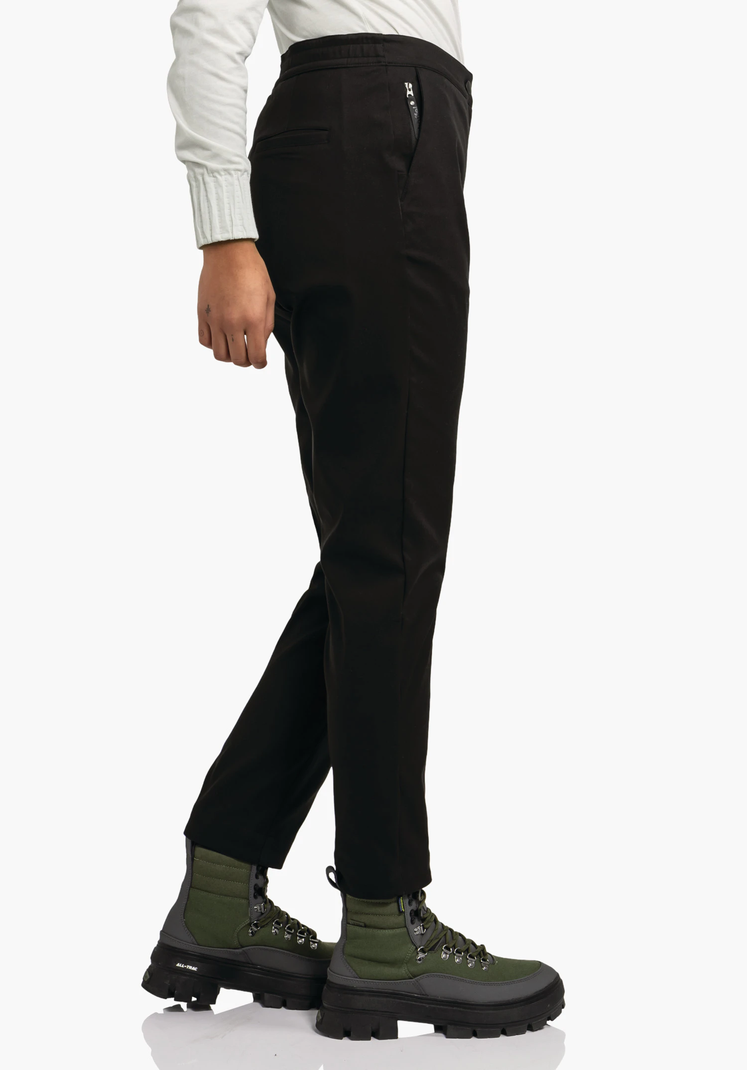 Pants Style Zourga WMS zwarte broek, zijwaarts gedragen met licht gekruist been; hand ontspannen langs het lichaam; context: witte achtergrond; lichtwit bovenstuk; donkergroene laarzen met zwarte zolen.