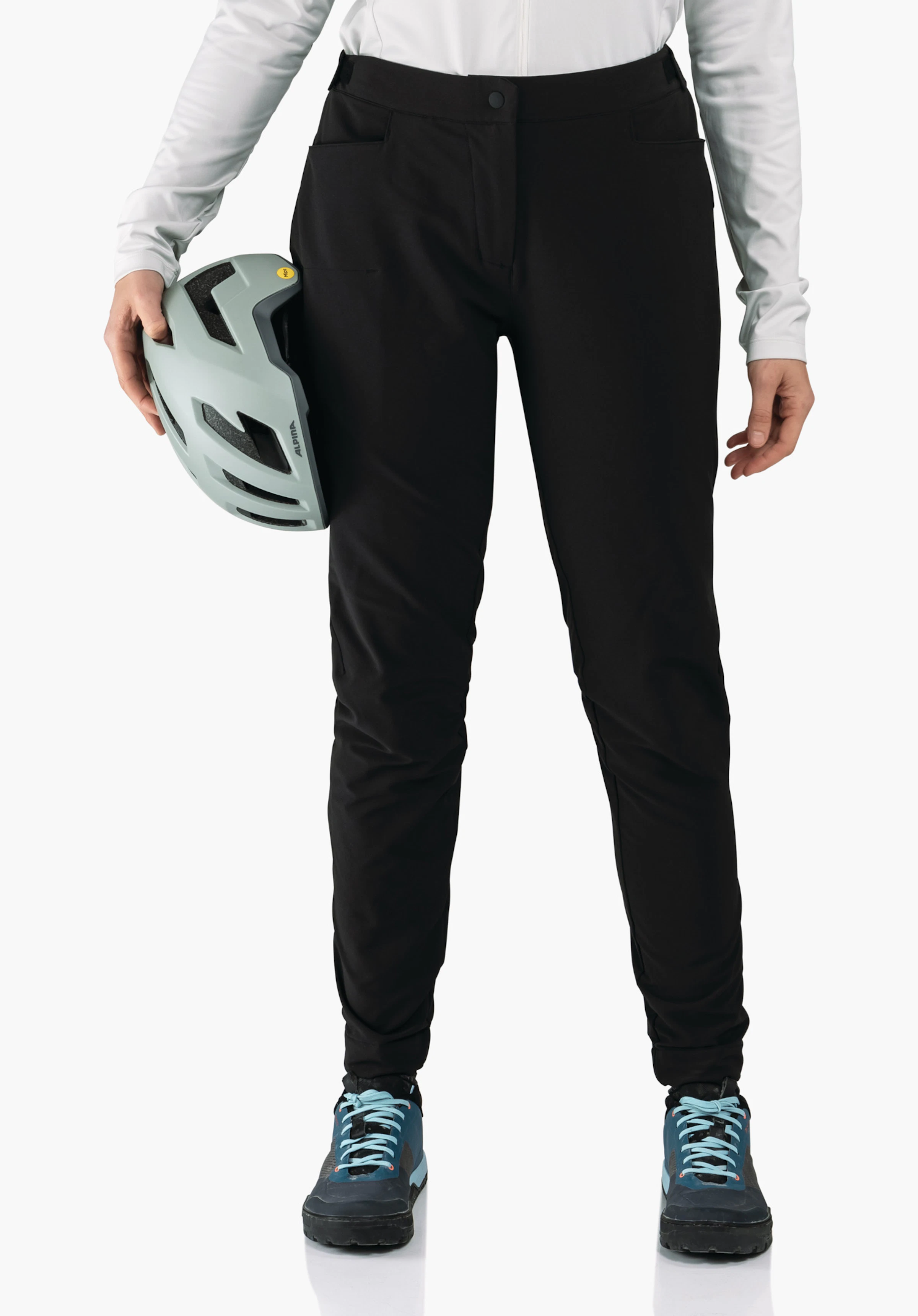 Pants Style Kubena WMS pantalon noir; personne tenant un casque de vélo gris clair avec inscription ALPINA, portant un haut manches longues blanc et des chaussures bleu foncé devant un fond clair uni.