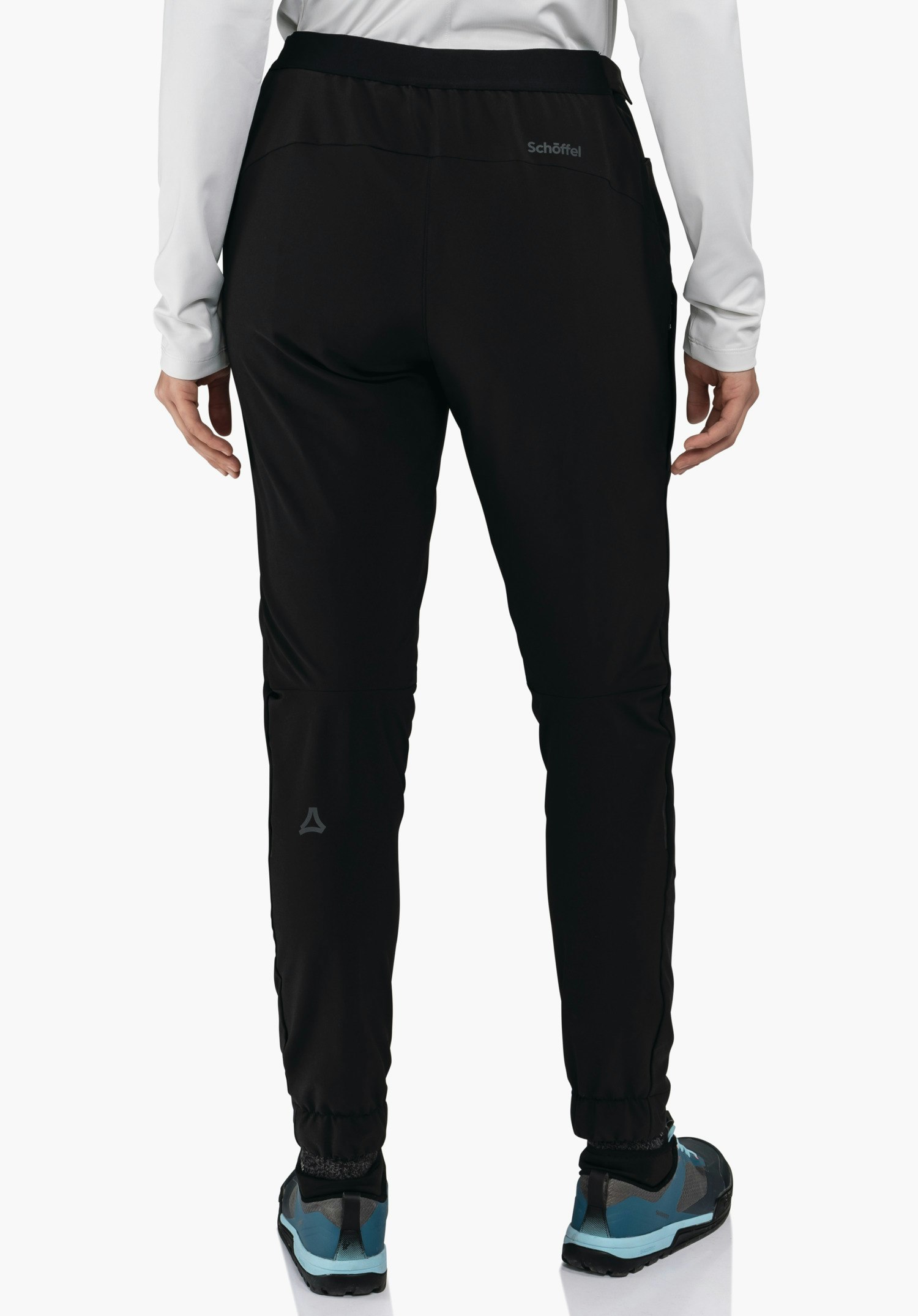 Pants Style Kubena WMS zwarte broek, gedragen door een persoon in achteraanzicht; witte top, donkerblauw-lichtblauwe wandelschoenen, Schöffel-logo op de kuit, opschrift "Schöffel" aan de tailleband.