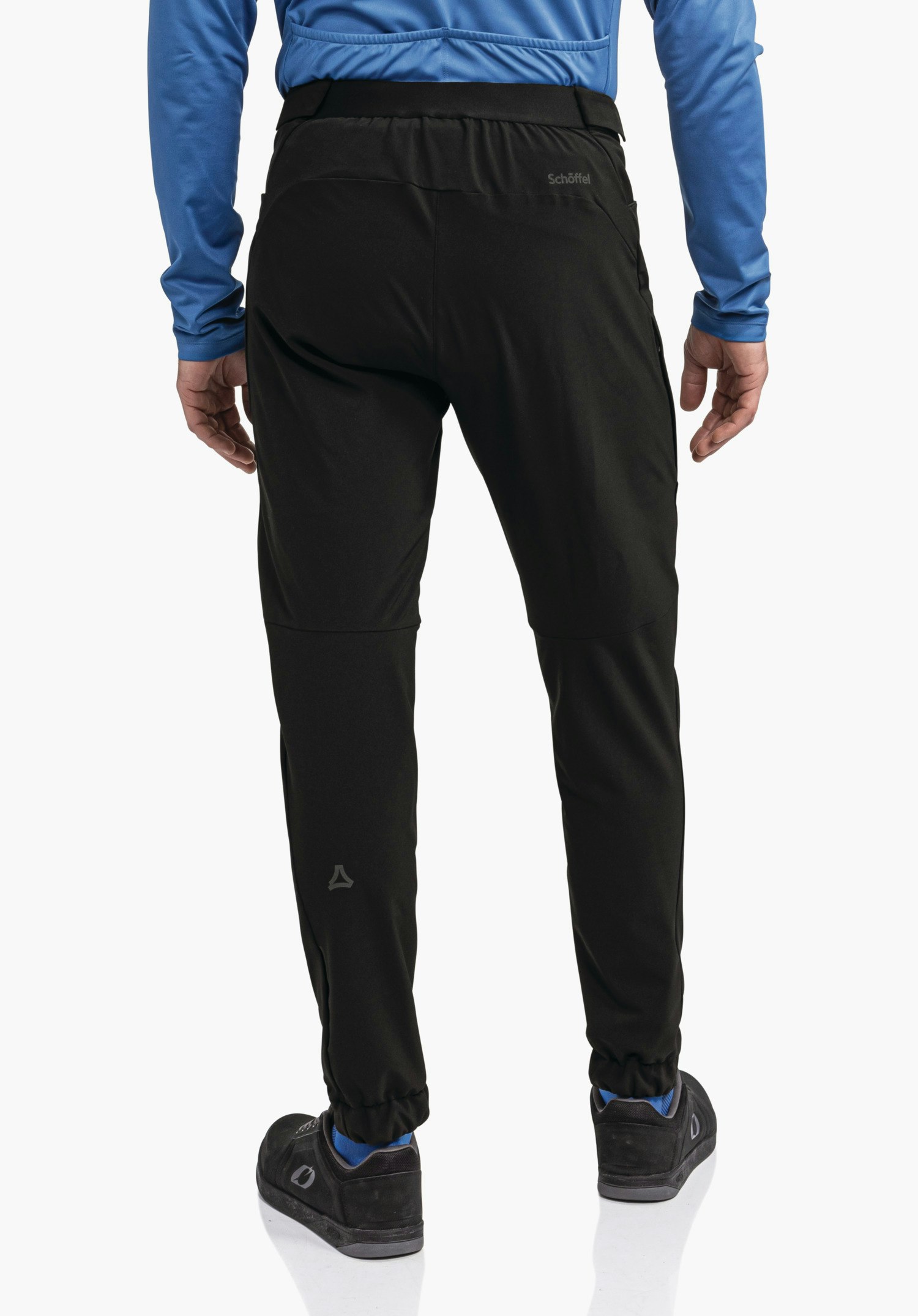Pants Style Kubena MNS black training trousers worn standing, rear view; “Schöffel” text at the upper right waistband, Schöffel logo on the left calf; paired with a dark blue long-sleeve top; white background.