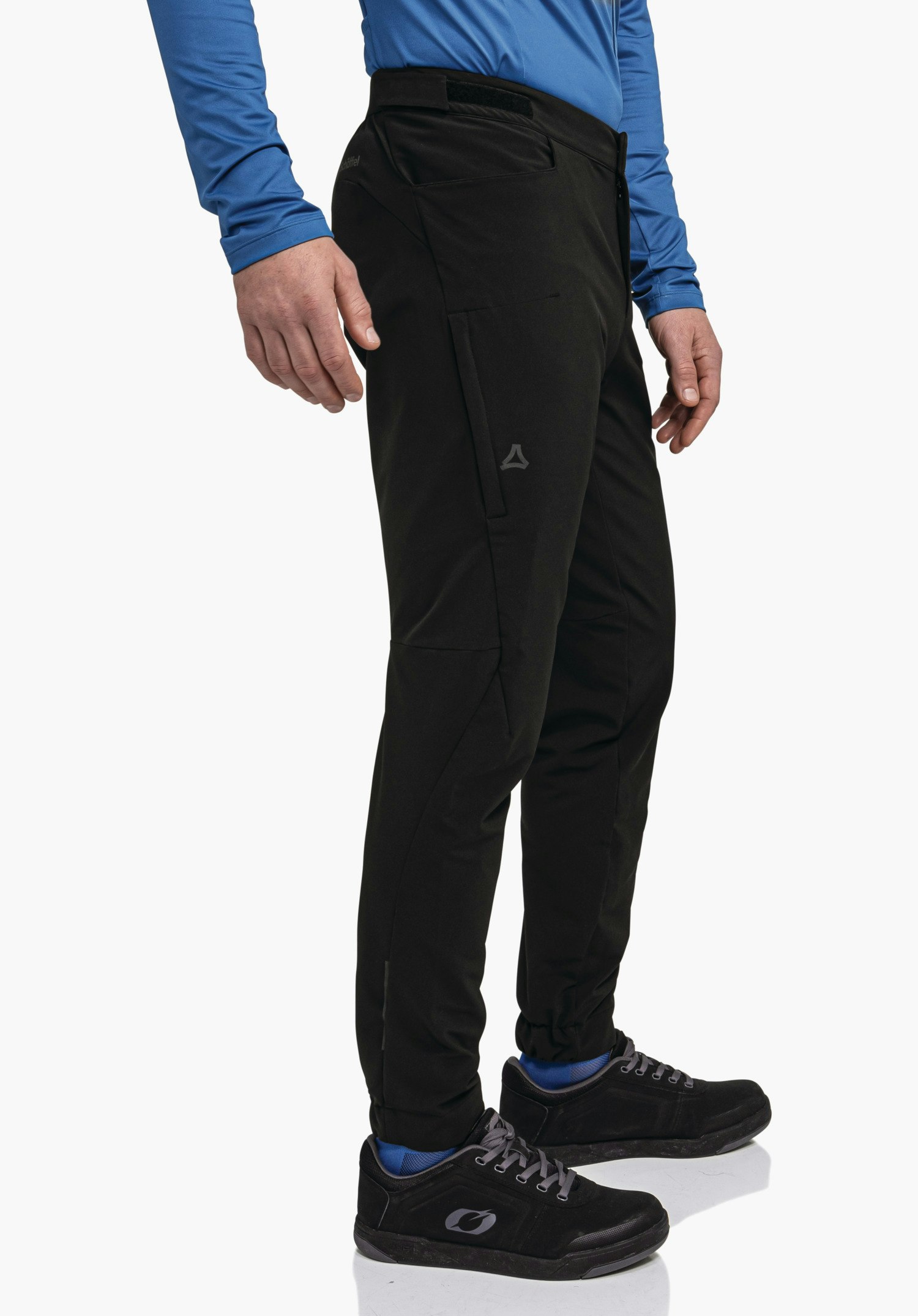 Pants Style Kubena MNS black outdoor trousers with Schöffel logo, worn side-on by a person; person stands slightly angled, blue top, blue socks, black shoes in front of a white background.
