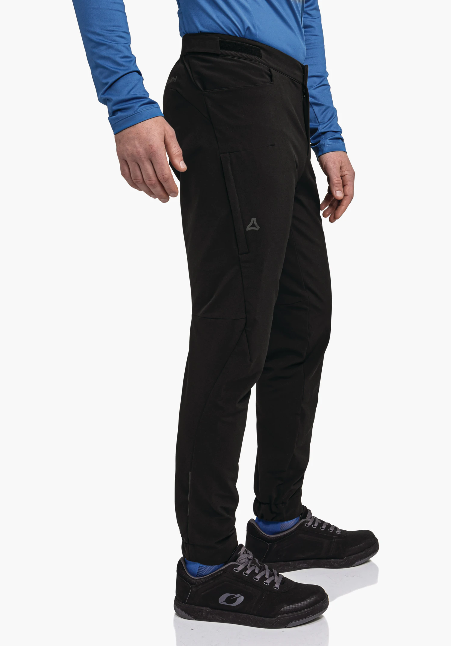 Pants Style Kubena MNS black outdoor trousers with Schöffel logo, worn side-on by a person; person stands slightly angled, blue top, blue socks, black shoes in front of a white background.