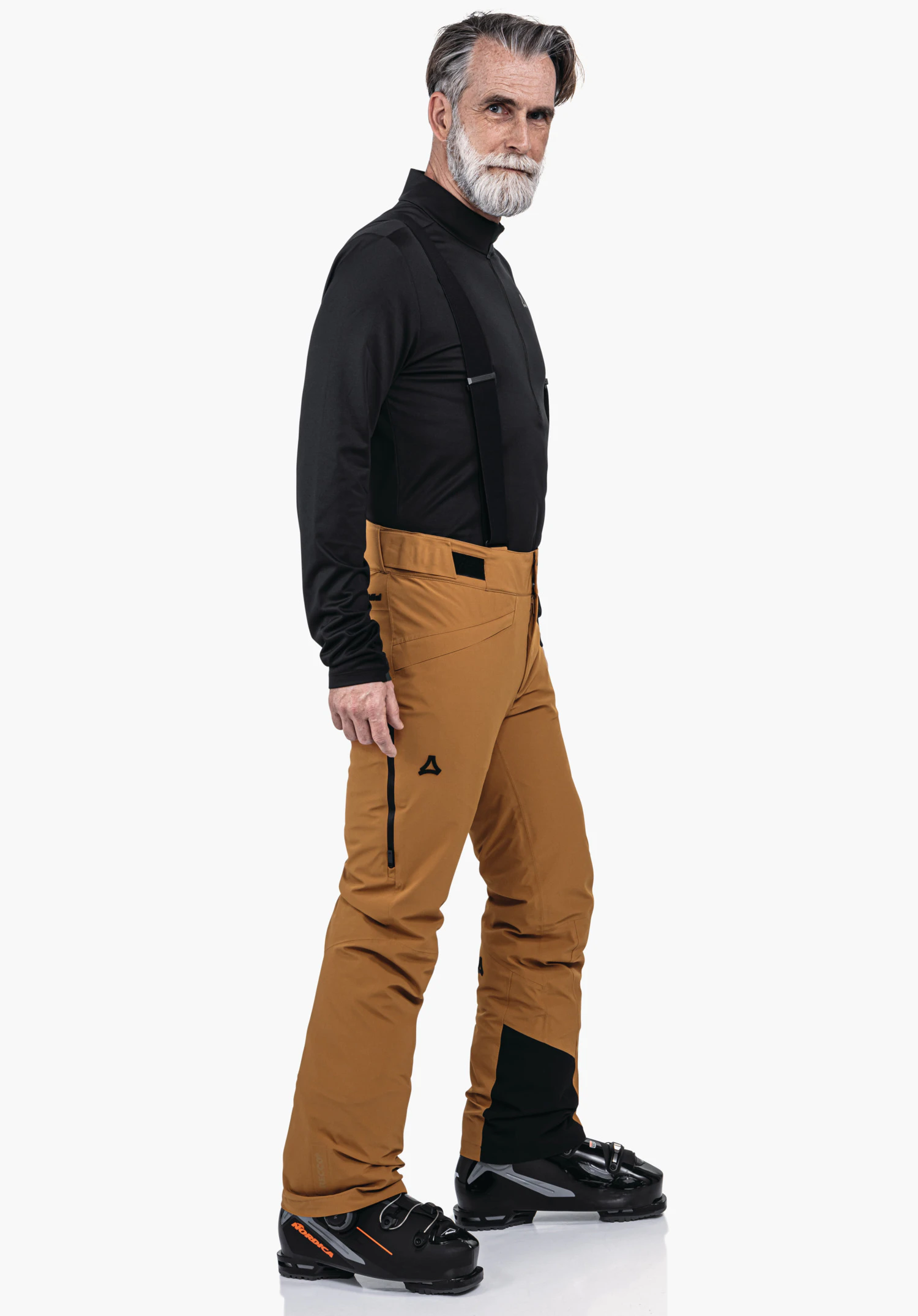 Pants Style Mountet MNS man stands side-on wearing a black long-sleeve top, orange ski trousers with Schöffel logo and black ski boots; looks ahead in front of a neutral white background.