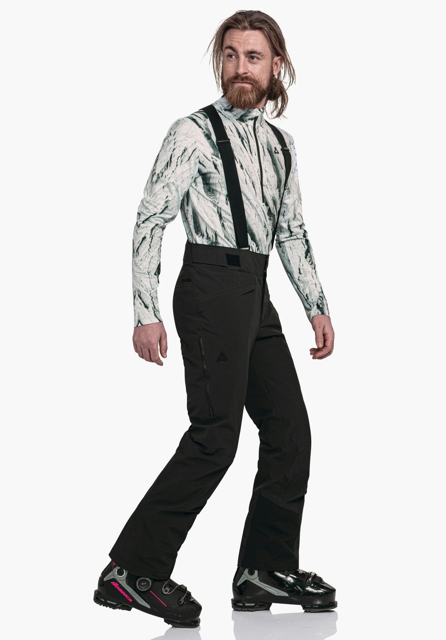Pants Style Mountet MNS Mann trägt ein hellweißes Oberteil mit dunkelgrünem Muster und schwarze Skihose mit Schöffel-Logo, geht seitlich und blickt nach rechts, vor weißem Studiohintergrund.