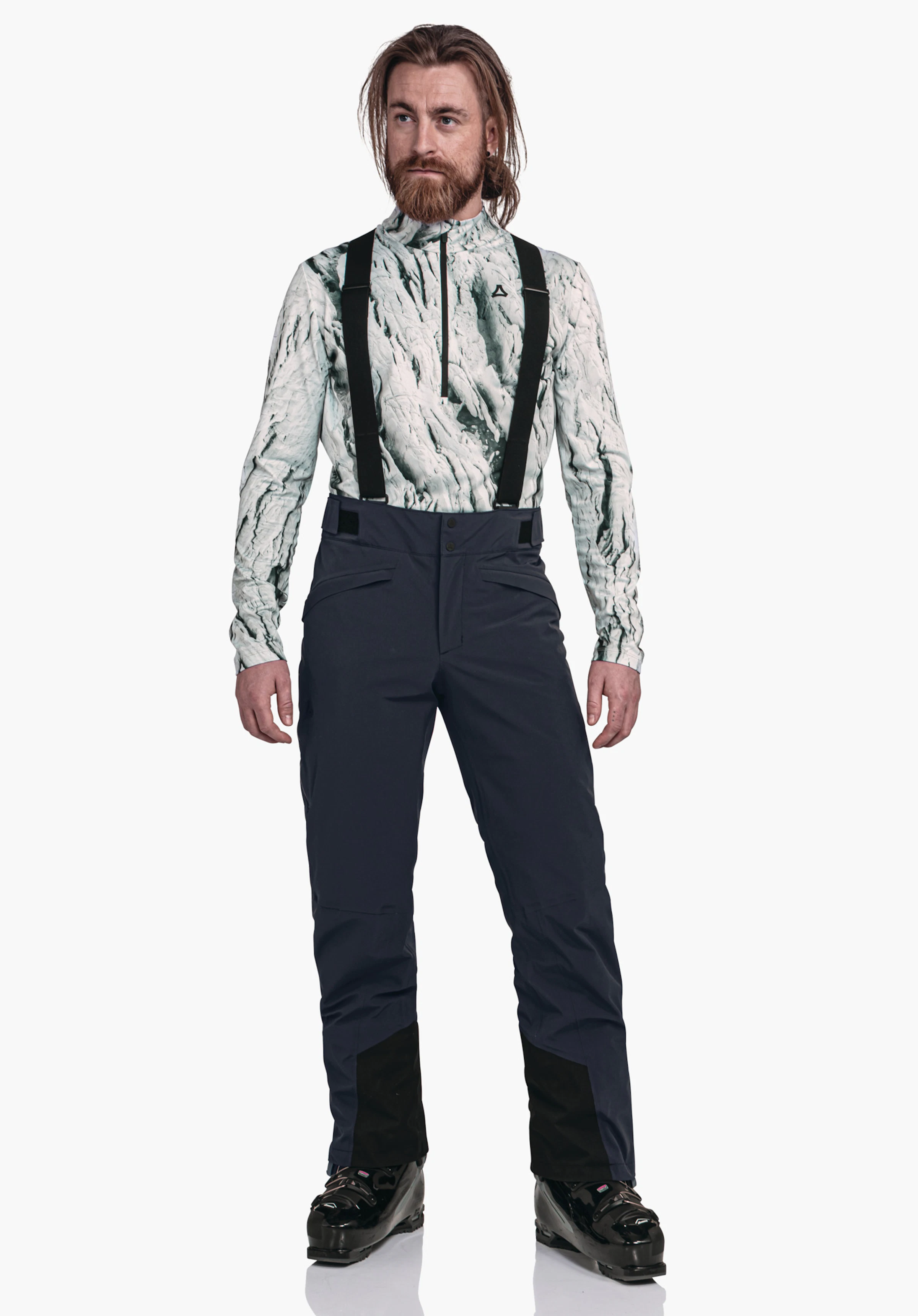 Pants Style Mountet MNS Mann trägt Skioutfit mit Schöffel-Logo, steht aufrecht, blickt seitlich; weiß gemustertes Oberteil, dunkelblaue Skihose mit schwarzen Hosenträgern, heller Studiohintergrund.