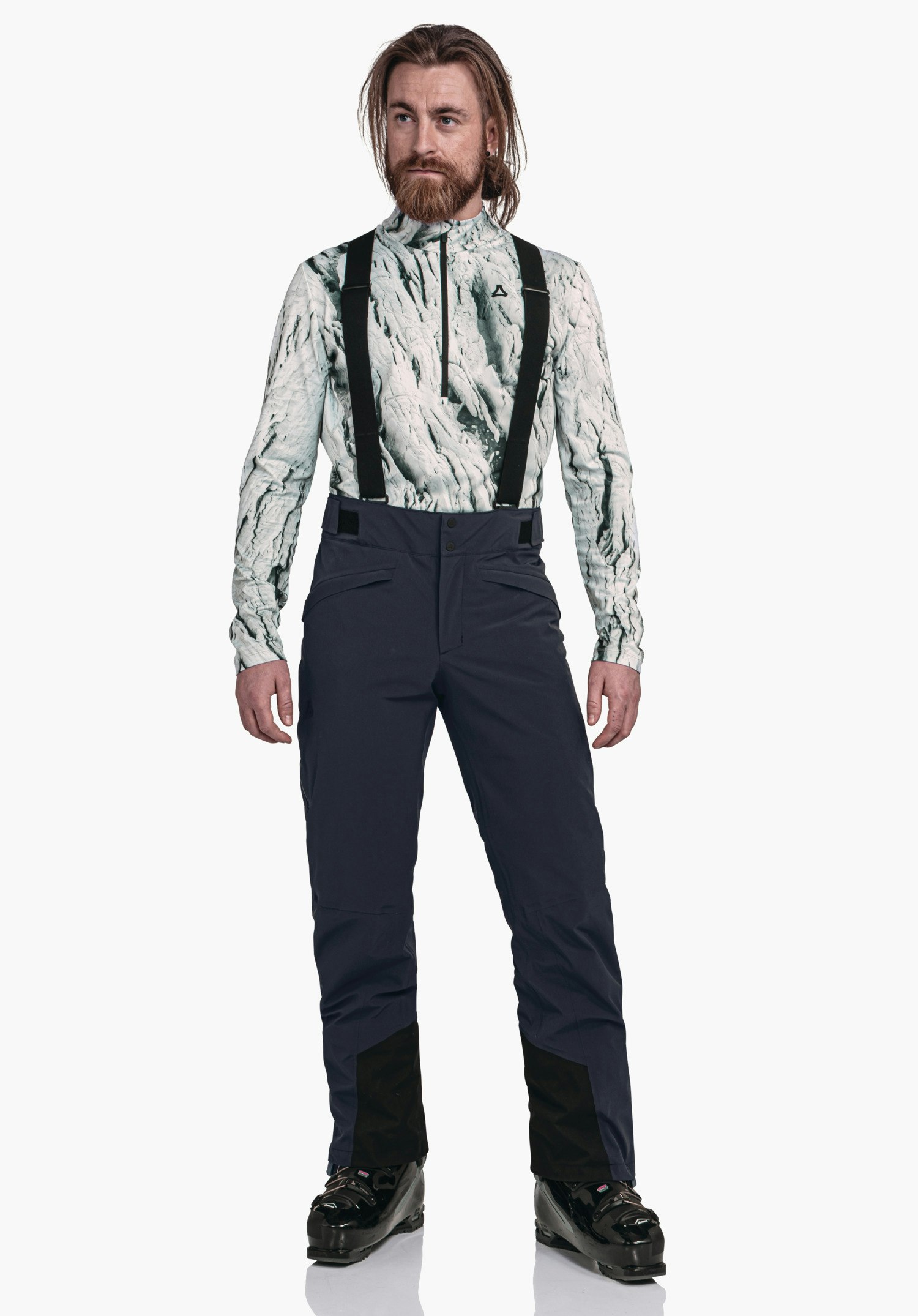 Pants Style Mountet MNS man draagt een ski-outfit met Schöffel-logo, staat rechtop en kijkt zijwaarts; wit gemêleerde top, donkerblauwe skibroek met zwarte bretels, lichte studioachtergrond.