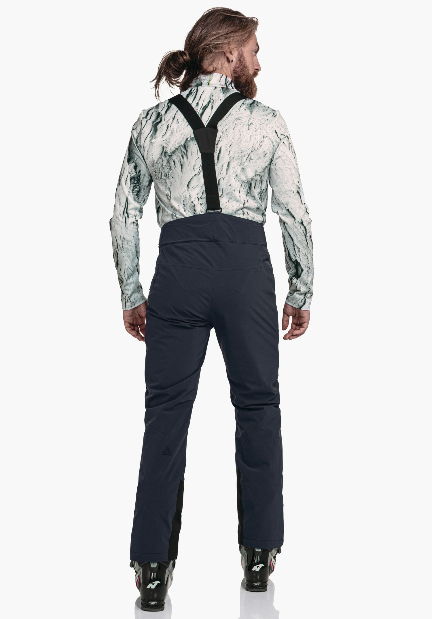 Pants Style Mountet MNS man in donkerblauwe skihose met zwarte bretels staat achterwaarts; lichtwit gemêleerd shirt, bruin haar in paardenstaart, zwarte skischoenen; Schöffel-logo links op het onderbeen, lichte achtergrond.