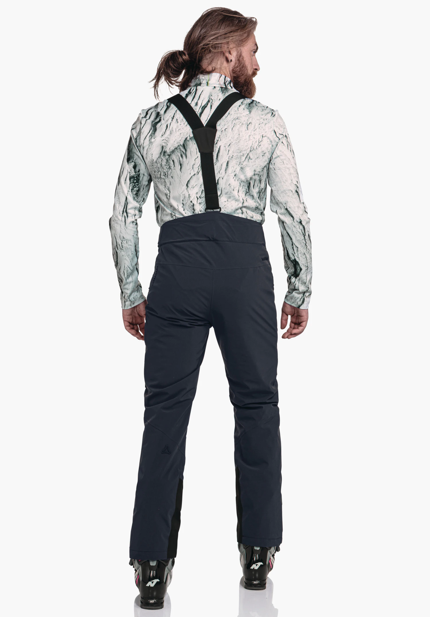 Pants Style Mountet MNS man wearing dark blue ski trousers with black braces, standing with back to the camera; off-white patterned shirt, brown hair in a ponytail, black ski boots; Schöffel logo on the left lower leg; light background., Pants Style Mountet MNS man wearing dark blue ski trousers with black braces, standing with back to the camera; off-white patterned shirt, brown hair in a ponytail, black ski boots; Schöffel logo on the left lower leg; light background.