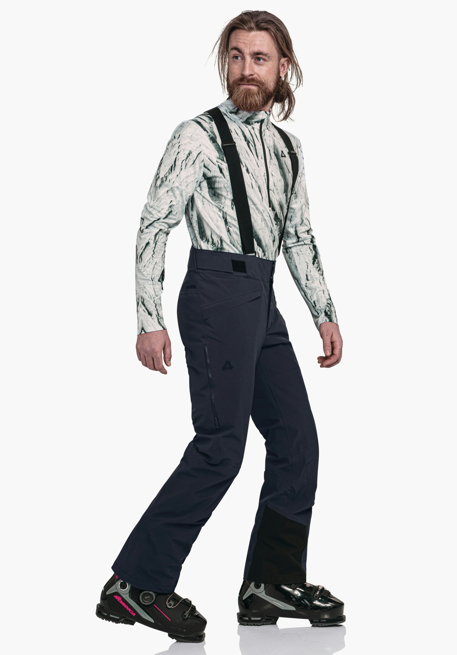 Pants Style Mountet MNS man draagt een lichtwit bovenstuk met donkergroen patroon en donkerblauwe skibroek met zwarte bretels; Schöffel-logo zichtbaar; loopt zijwaarts voor een witte studioachtergrond, zwarte skischoenen met rode details.