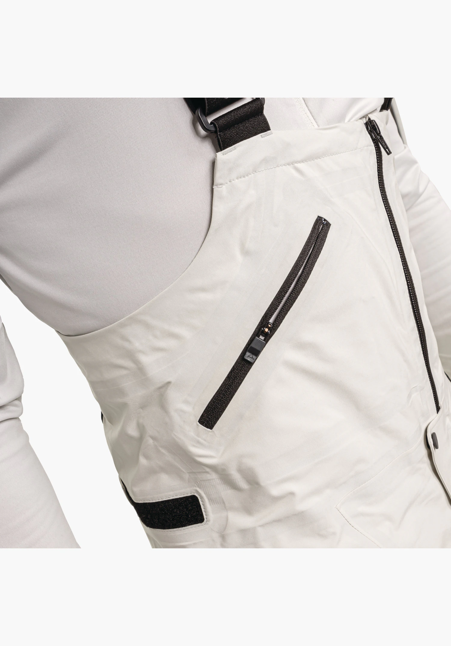 Pants Style Lufeld MNS detail view of the bib in nature colour featuring a diagonal zip pocket, adjustable strap and side fastening, close-up against a white/neutral background.