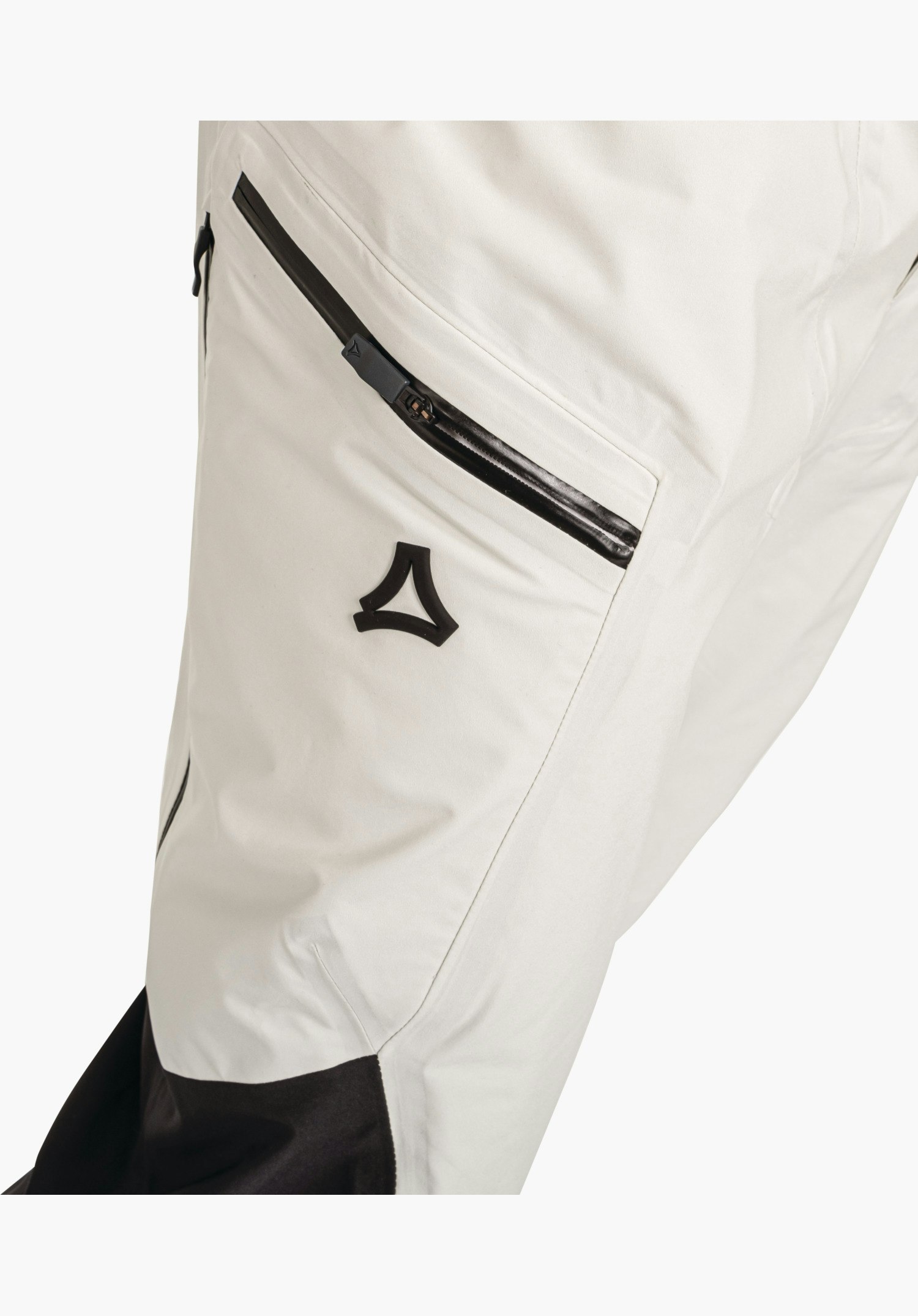 Pants Style Lufeld MNS toont de contrasterende zwarte rits van de dijbeenzak en het opliggende Schöffel-logo op de natuurkleurige stof. Close-up tegen een witte/neutrale achtergrond.