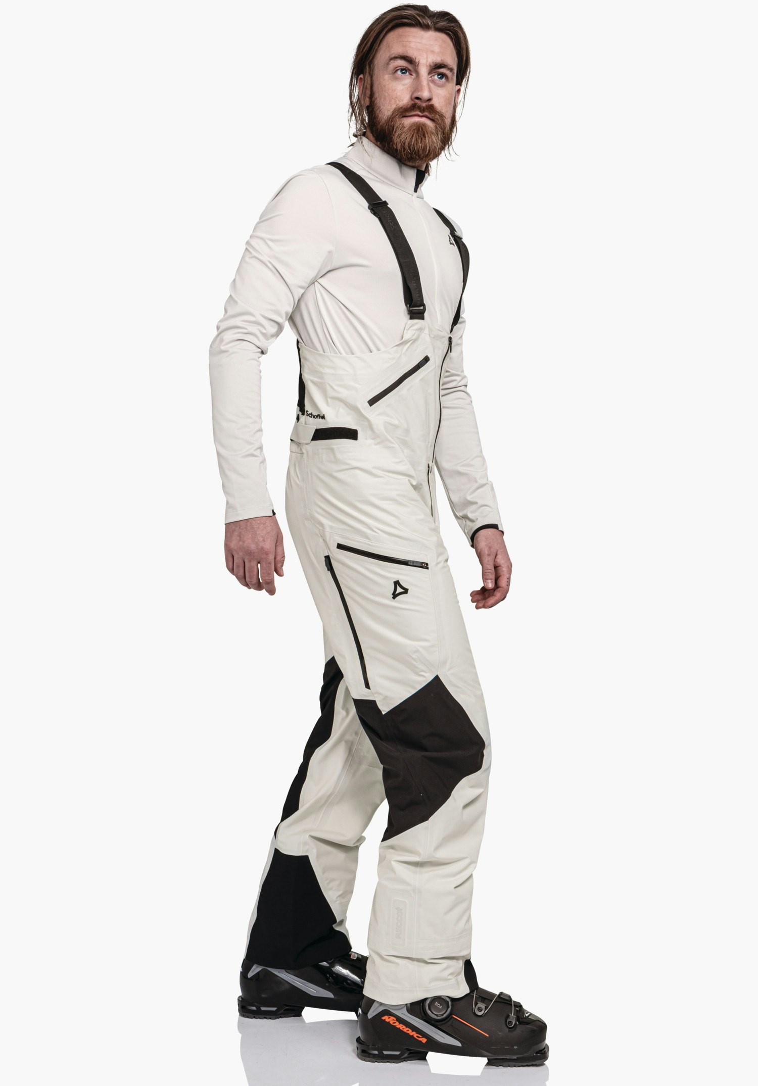 Pants Style Lufeld MNS light white ski trousers with black insert, bib with braces; man stands mid-step, studio in front of a white background. Text: Schöffel, Schöffel logo, NORDICA.