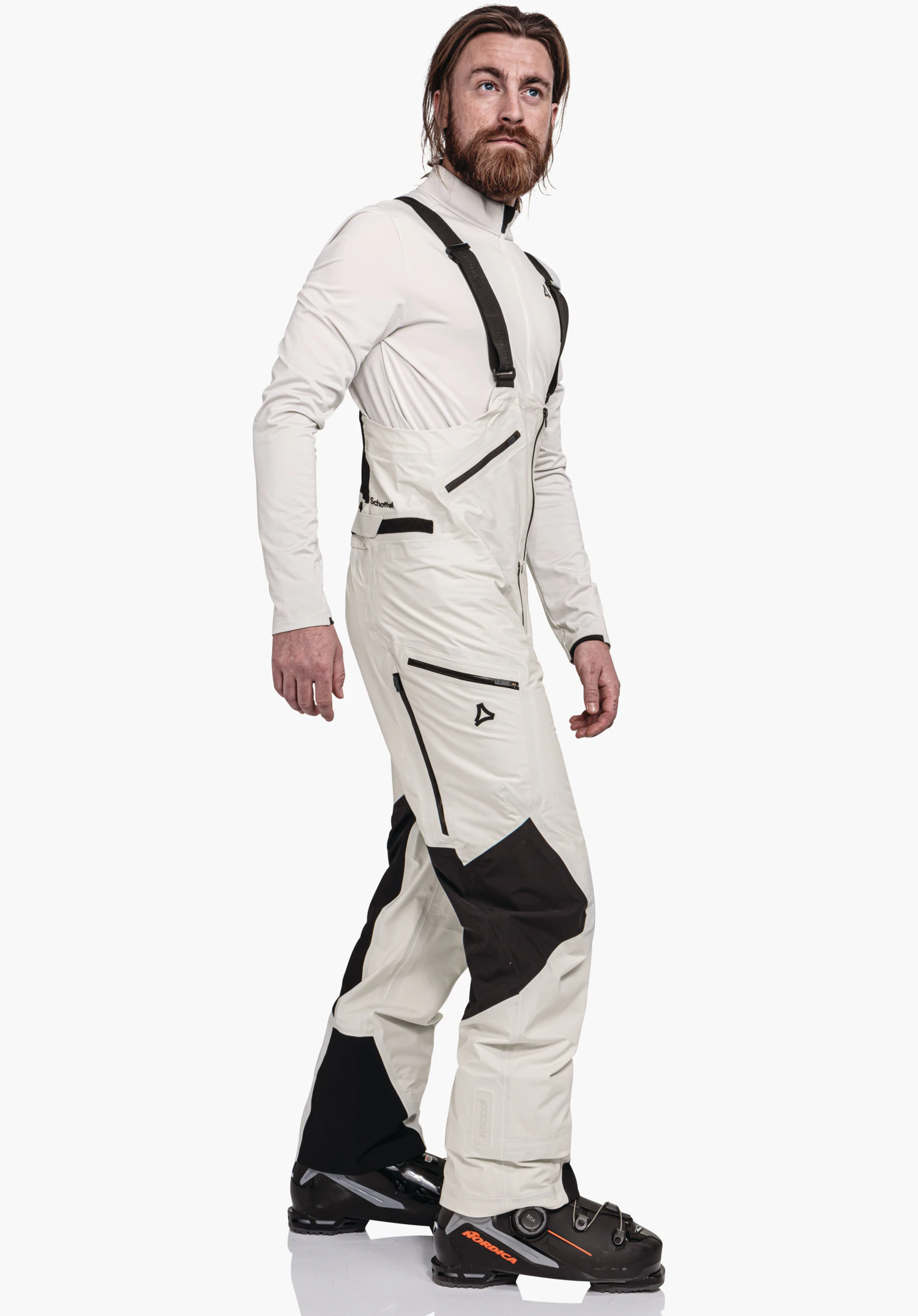 Pants Style Lufeld MNS light white ski trousers with black insert, bib with braces; man stands mid-step, studio in front of a white background. Text: Schöffel, Schöffel logo, NORDICA.