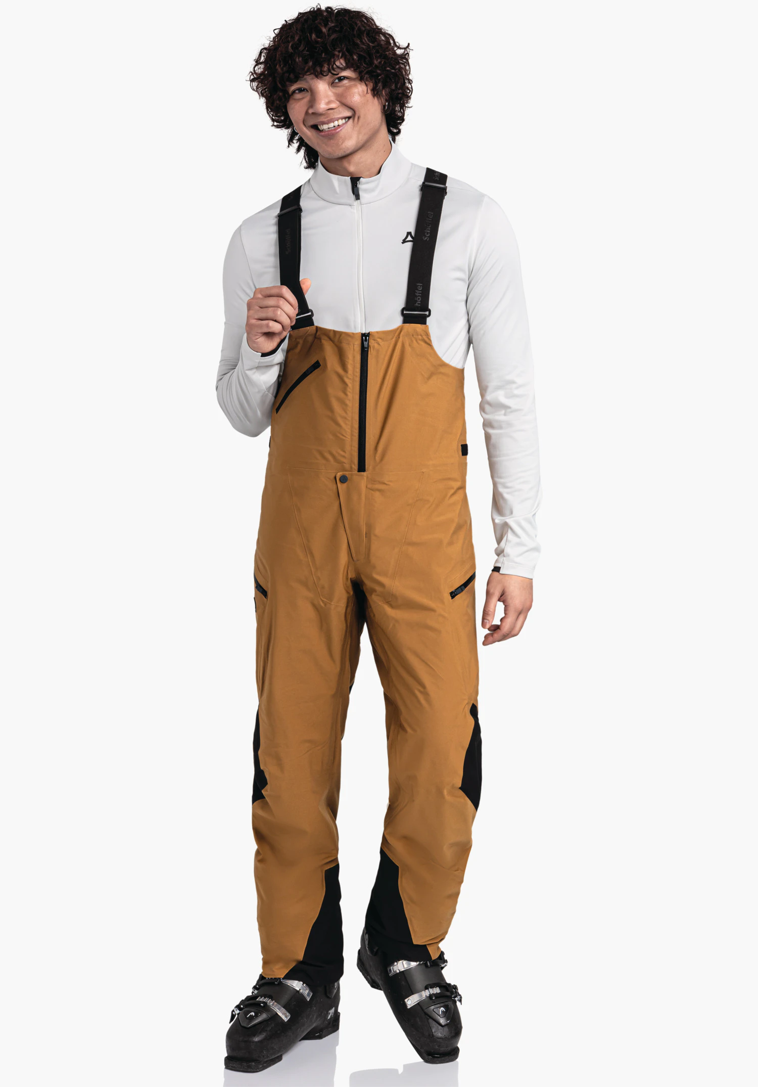 Pants Style Lufeld MNS Mann trägt orange Latzhose mit schwarzen Trägern; hält einen Träger. Weißes Oberteil mit Schöffel-Logo; steht vor weißem Hintergrund; schwarze Skischuhe. Text auf Trägern: "Schöffel".
