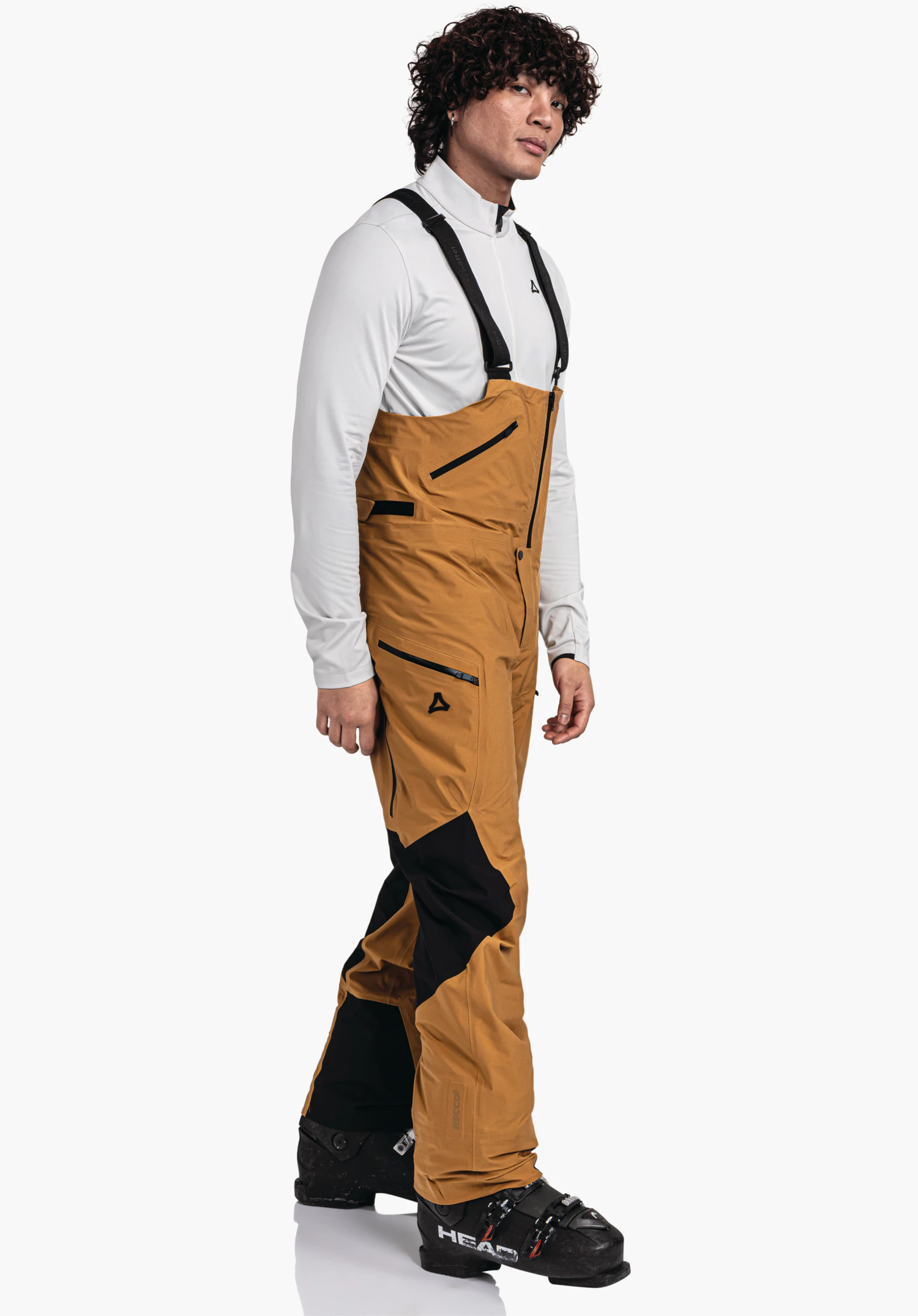 Pants Style Lufeld MNS dunkelgelbe Latzhose mit schwarzen Einsätzen und Schöffel-Logo; an einer stehenden Person getragen, seitlich gedreht, mit weißem Oberteil und schwarzen Skischuhen vor weißem Studiohintergrund.