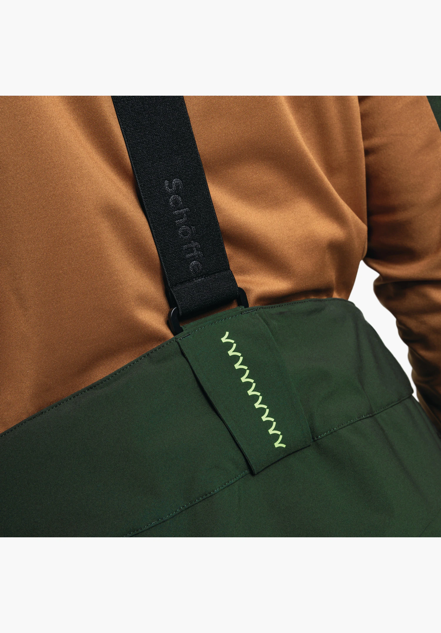 Pants Style Pine MNS avec un focus sur les bretelles noires avec inscription Schöffel et la ceinture verte avec éléments de design. Gros plan sur fond blanc/neutre.