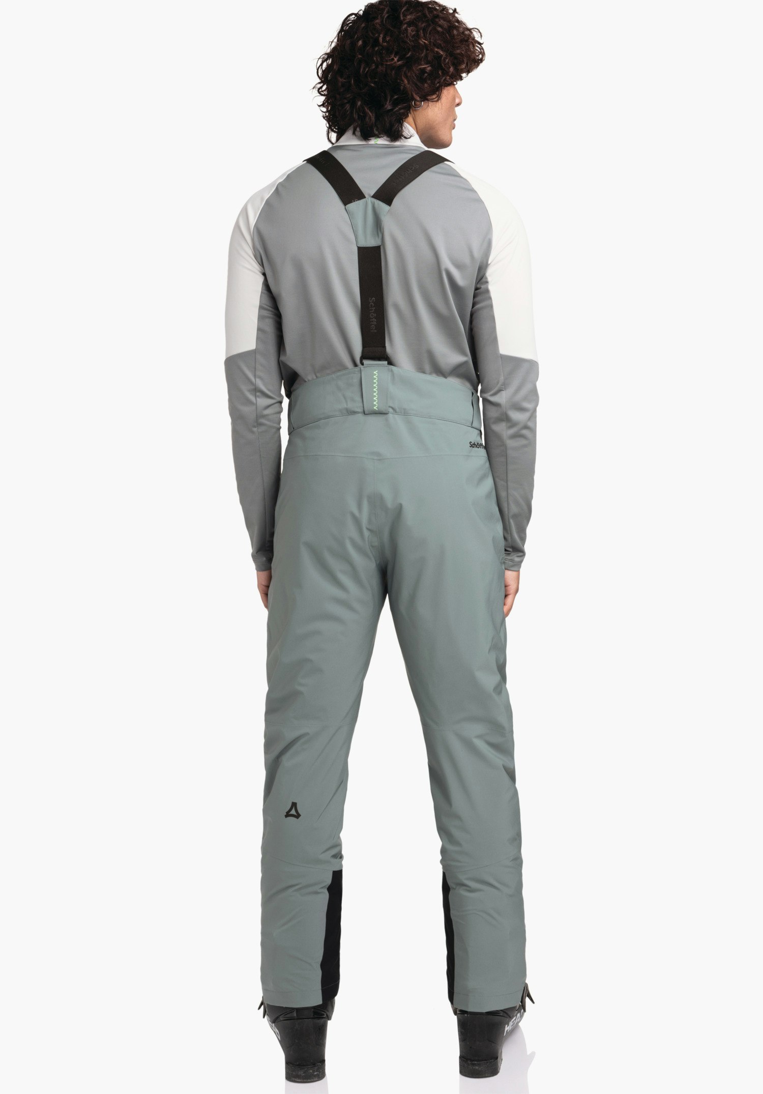 Pants Style Pine MNS Hellgrüne Skihose mit schwarzen Hosenträgern; Person steht rückwärts auf Skischuhen vor hellem Hintergrund, trägt weißes/hellgraues Oberteil. Schöffel-Logo am linken Bein. Text: "Schöffel".