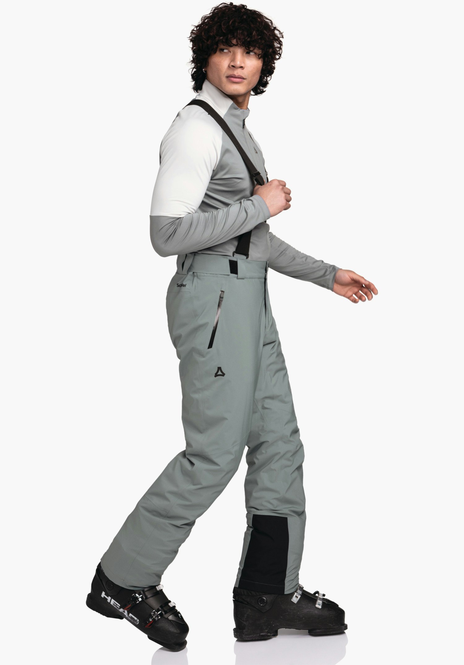 Pants Style Pine MNS Mann trägt hellgraue Skihose mit schwarzen Trägern, geht seitlich vor weißem Hintergrund, trägt weißes und hellgraues Oberteil; Schöffel-Logo auf Hose; Stiefel beschriftet: "HEAD".