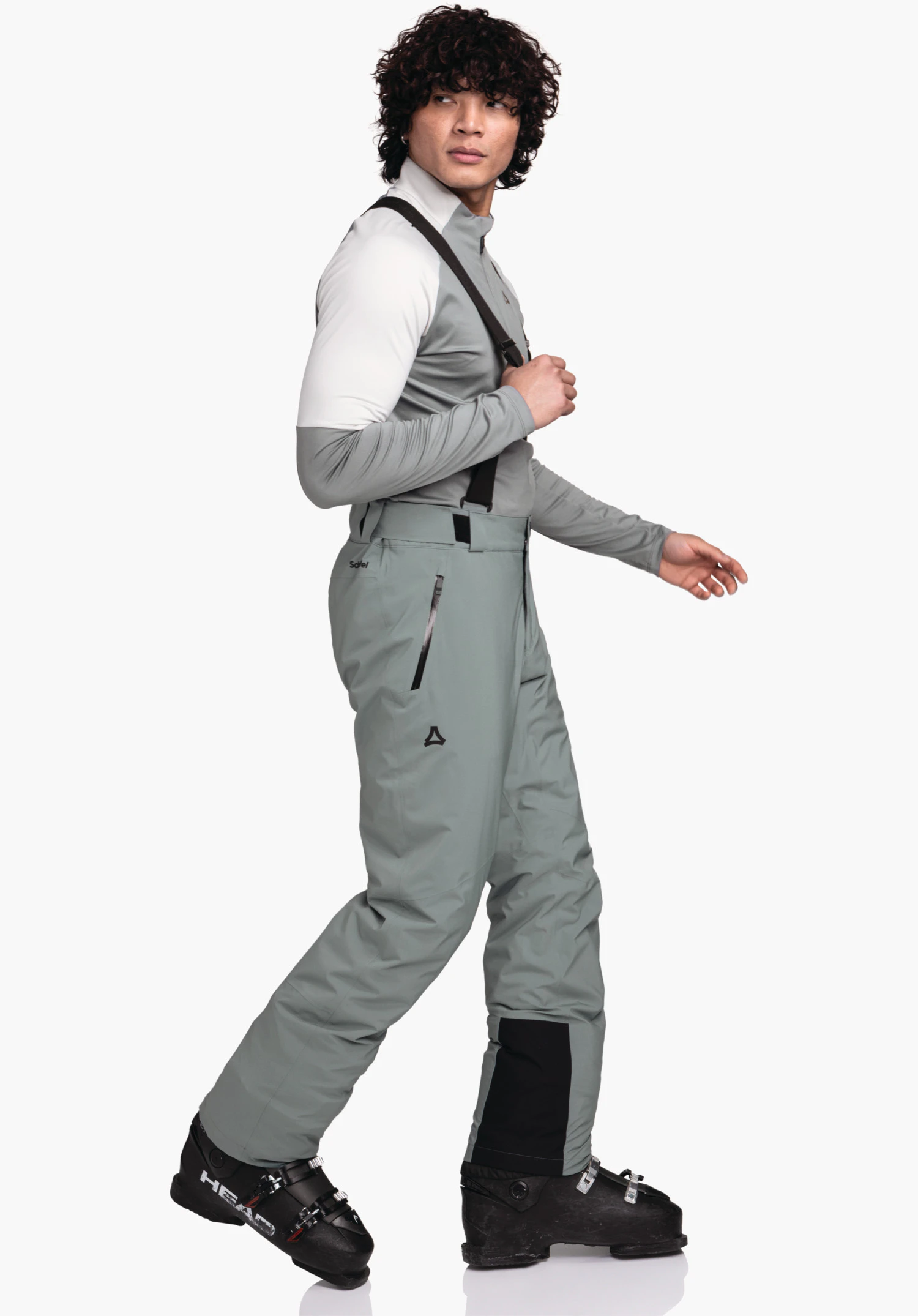Pants Style Pine MNS man wears light grey ski trousers with black braces, walks side-on in front of a white background, wears a white and light grey top; Schöffel logo on trousers; boots labelled: "HEAD".