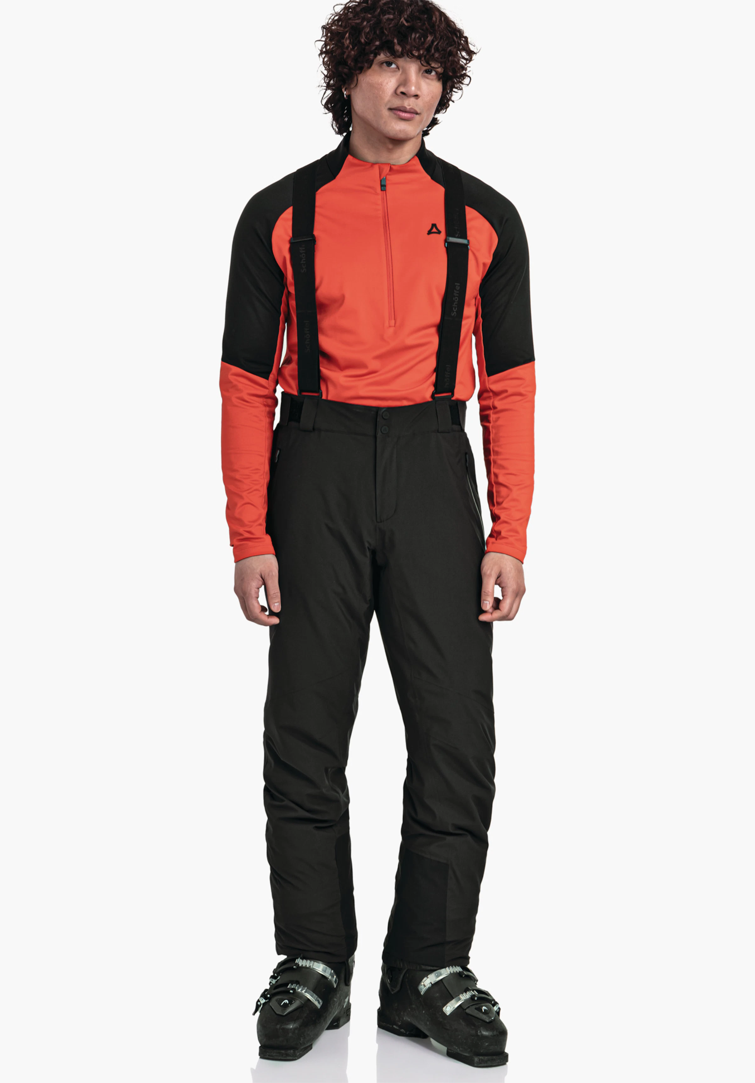 Pants Style Pine MNS Mann trägt ein Oberteil in hellrot mit Schöffel-Logo, hat schwarze Hosenträger an schwarzen Skihosen, steht in schwarz Skischuhen vor weißem Hintergrund. Text: "Schöffel".