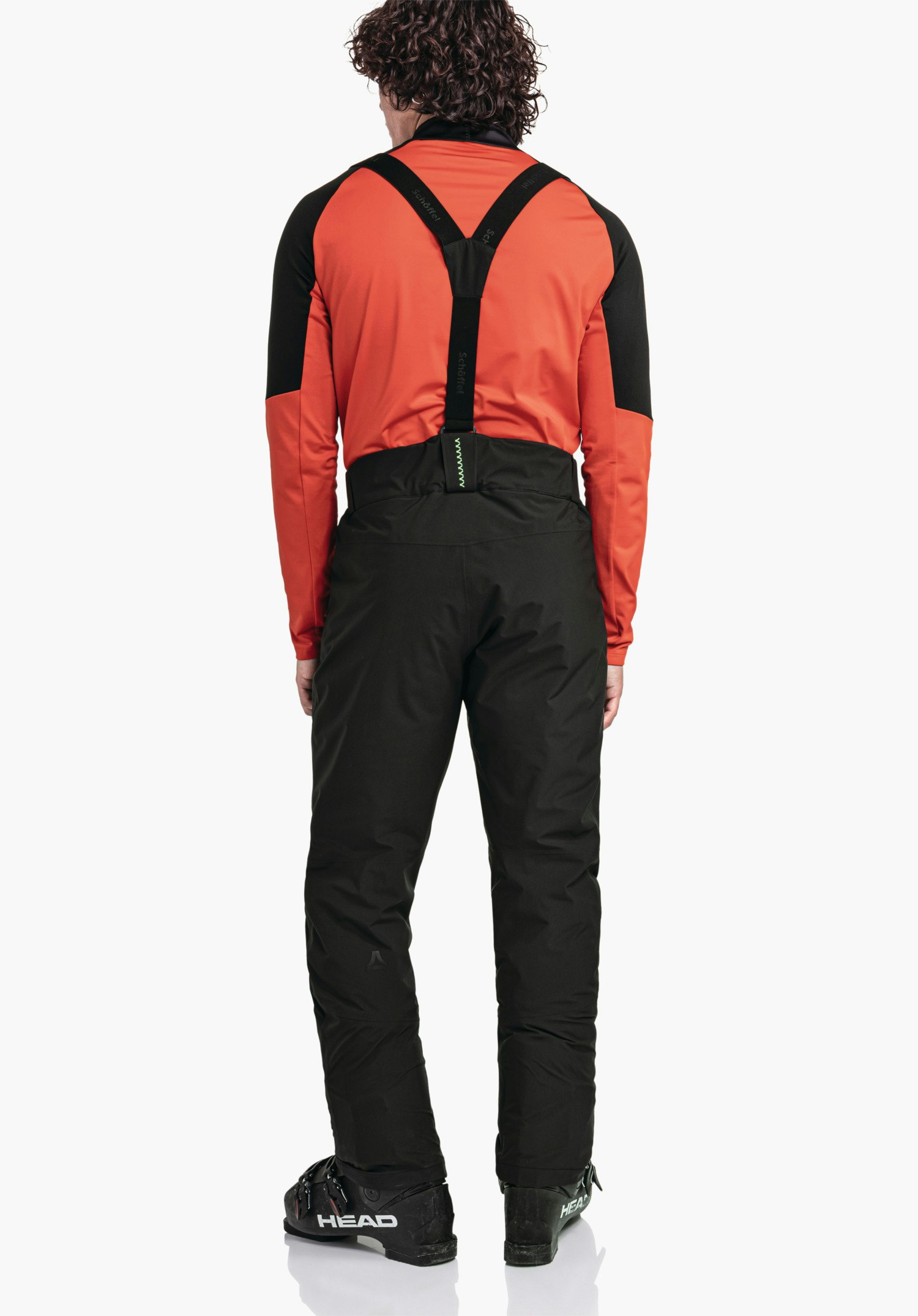 Pants Style Pine MNS persoon draagt een rode jas en een zwarte skihose met zwarte bretels en staat achterwaarts voor een witte achtergrond; Schöffel-logo zichtbaar. Tekst: "HEAD" op skischoenen, "Schöffel" op bretels.