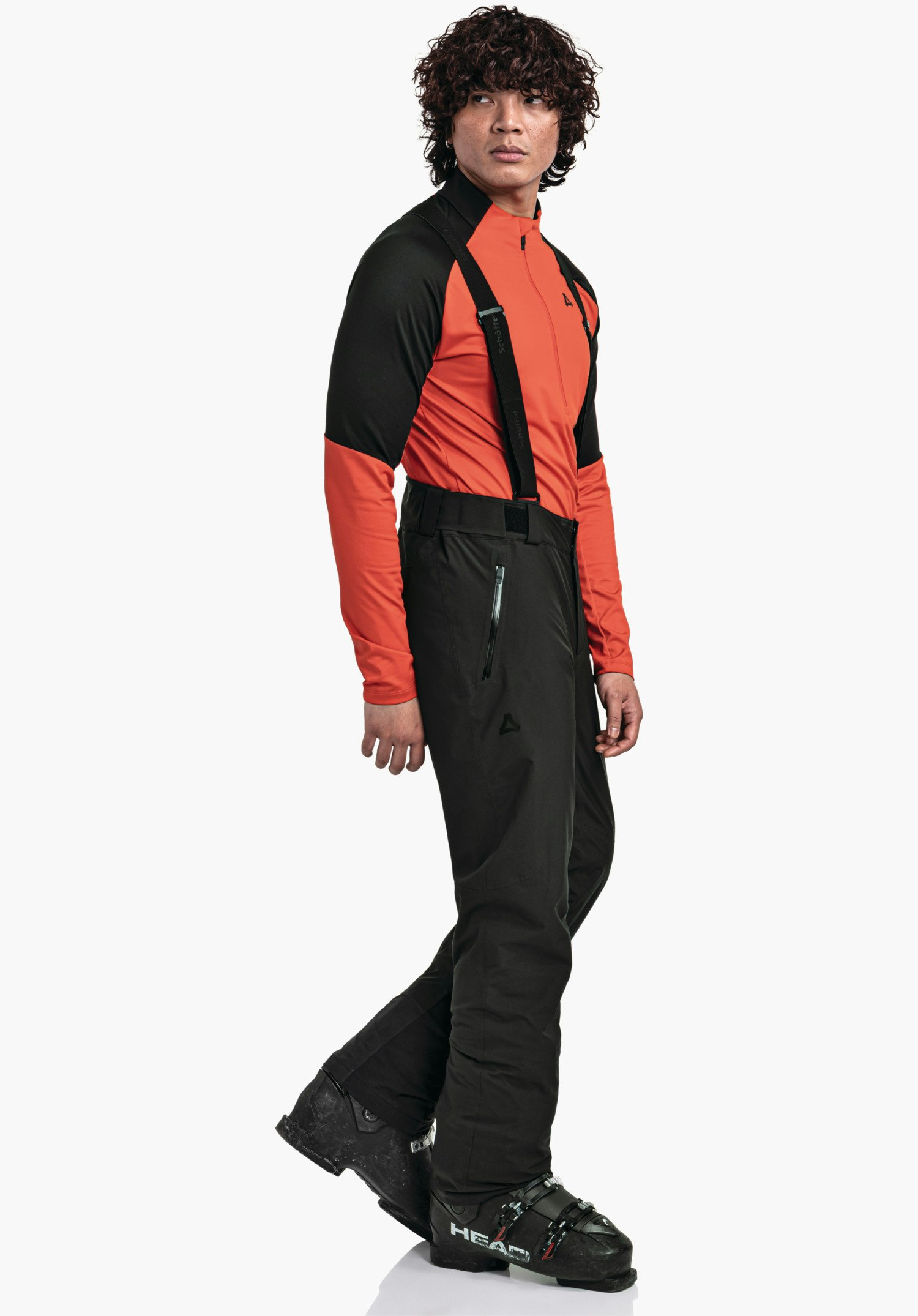 Pants Style Pine MNS man draagt rood-zwarte ski-kleding en een zwarte skibroek met bretels (Schöffel-logo), staat zijwaarts en kijkt naar rechts voor een witte studioachtergrond. Tekst: "HEAD".