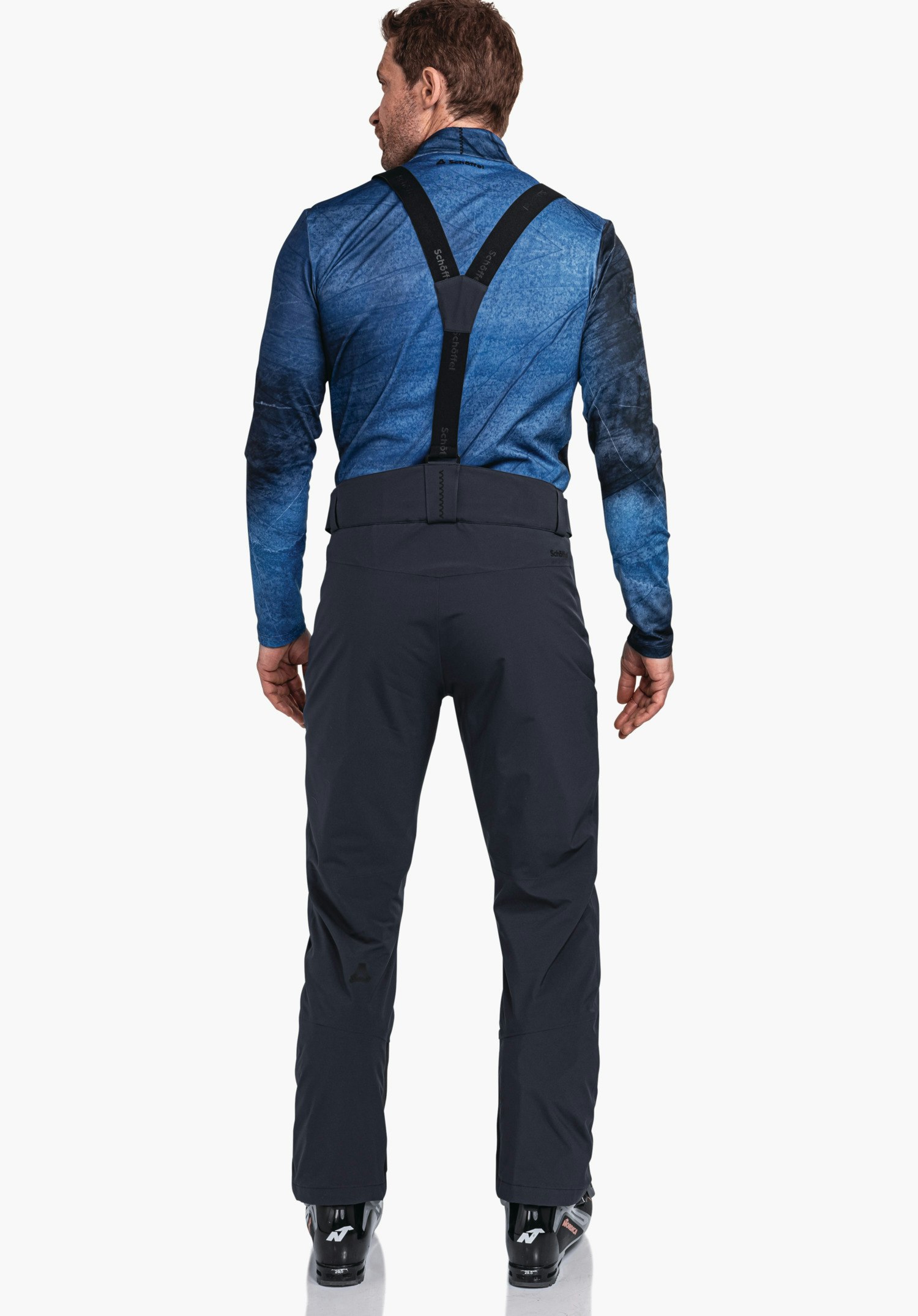 Pants Style Pine MNS Mann trägt dunkelblaues Langarmshirt und dunkelgraue Skihose mit schwarzen Hosenträgern, steht rückwärts vor hellem Hintergrund; Schöffel-Logo an linker Wade.  Text: "schöffel" (auf Hosenträgern und am Hosenbund).