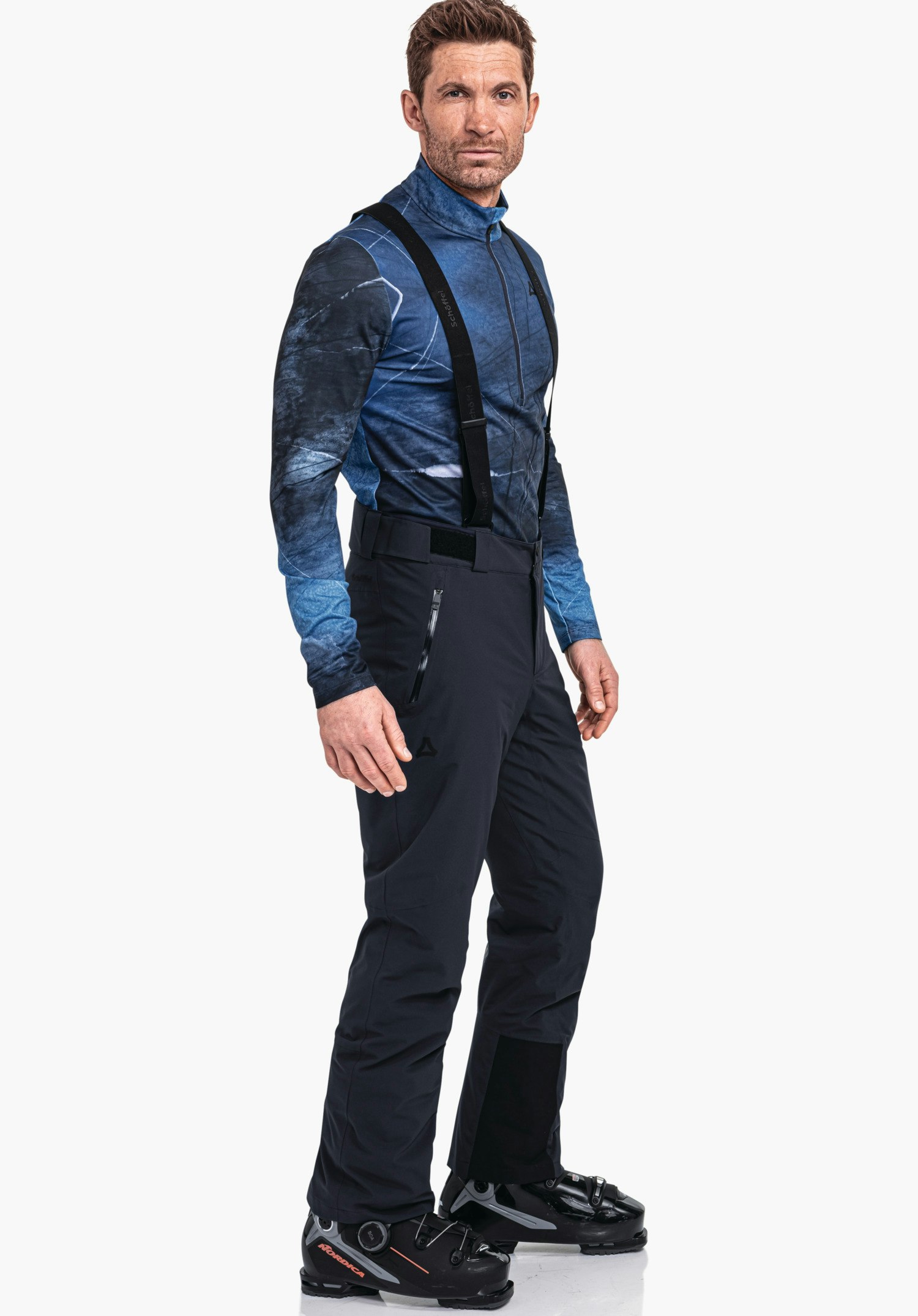 Pants Style Pine MNS Mann trägt hell blaues und dunkel blaues Skioberteil, dunkel blau Skihose mit Schöffel-Logo, steht leicht seitlich auf weißem Hintergrund; trägt schwarze Skischuhe mit orange Akzenten.