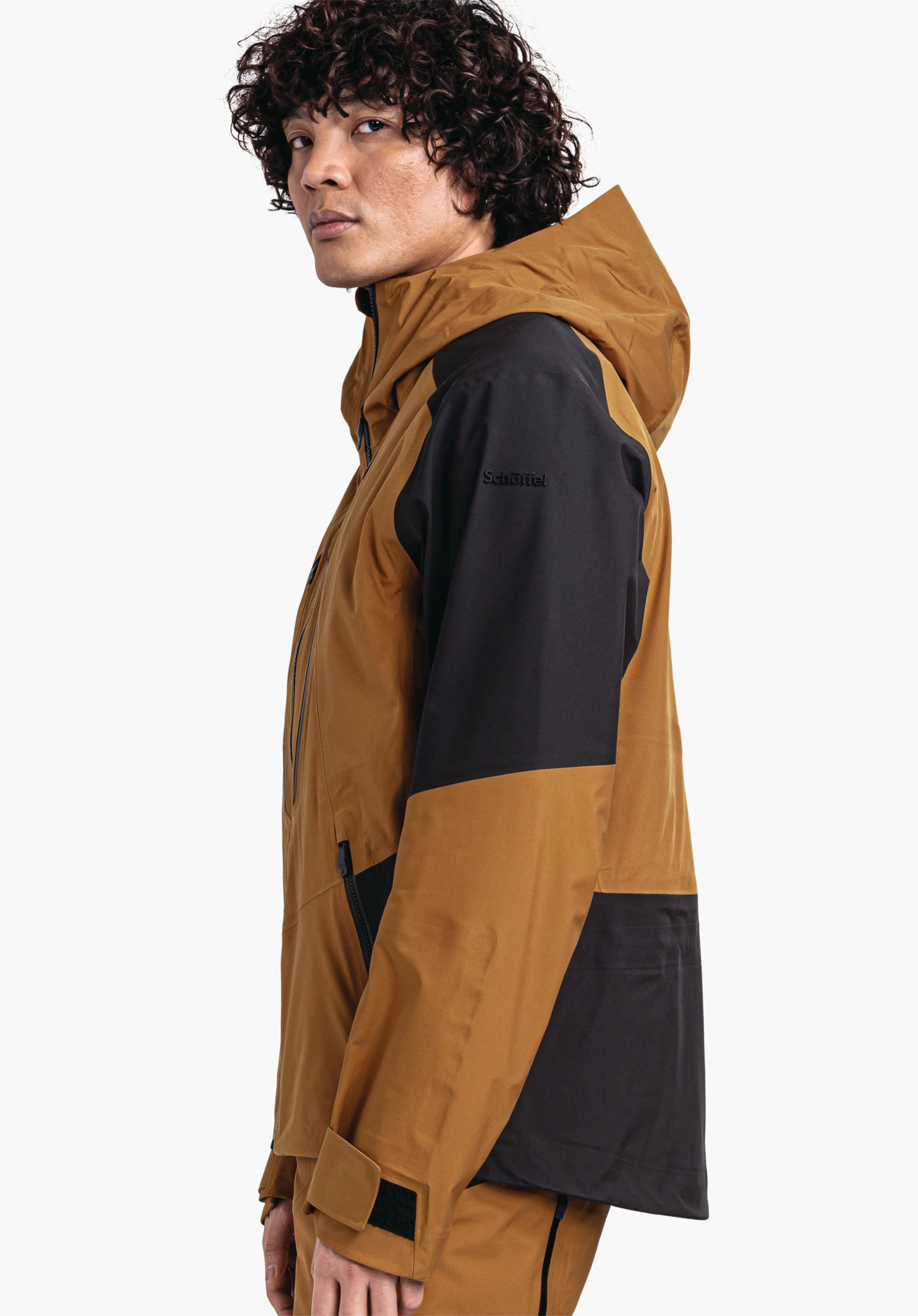 Shell Jk Style Lufeld MNS dunkelgelbe Jacke mit schwarzen Einsätzen, getragen von seitlich stehender Person, schaut zur Kamera vor weißem Studiohintergrund. Text auf Ärmel: "Schöffel".