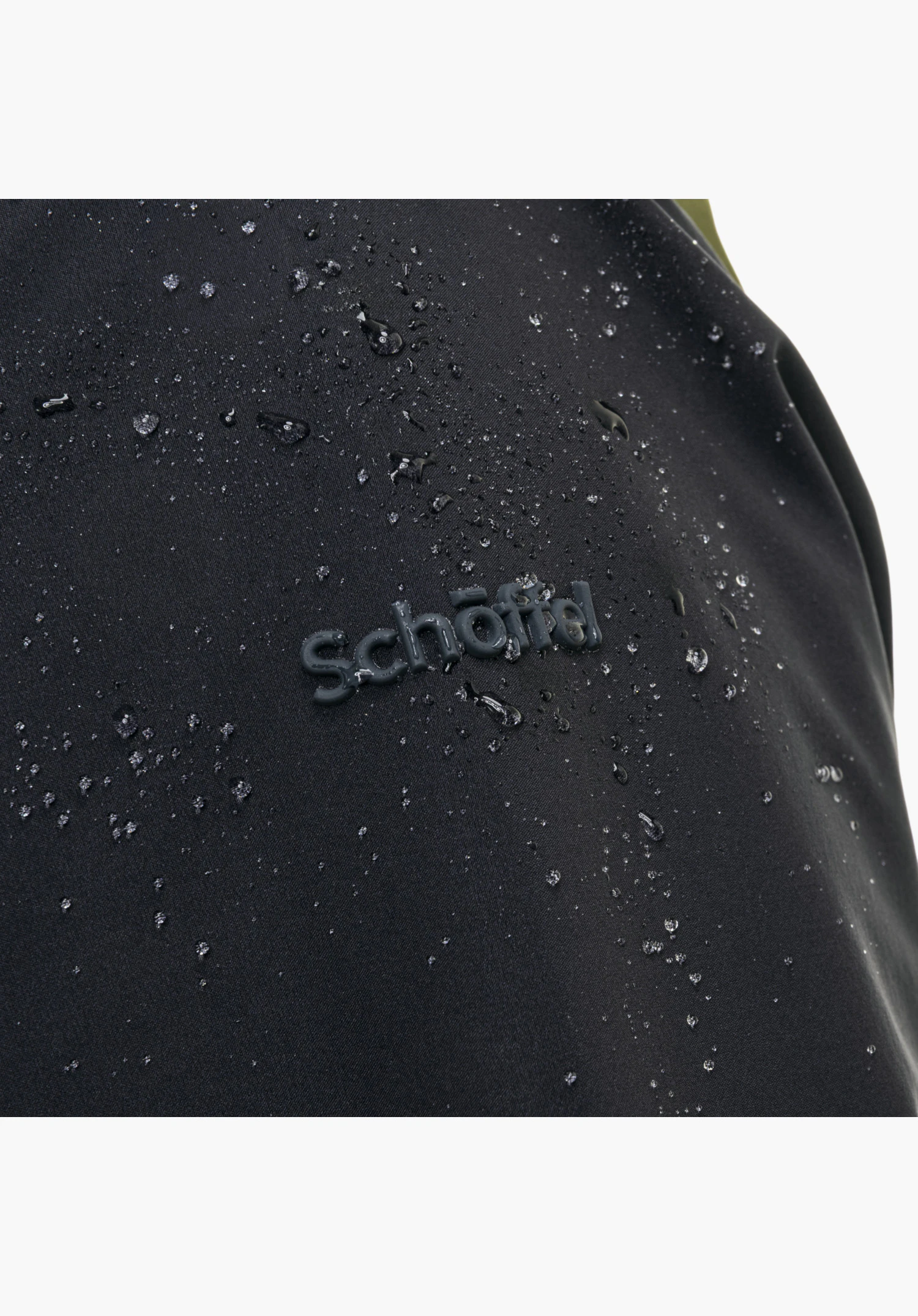 Shell Jk Style Lufeld MNS Detailaufnahme des grünen Materials mit dem Schöffel-Schriftzug und abperlenden Wassertropfen Nahaufnahme vor weißem Hintergrund.