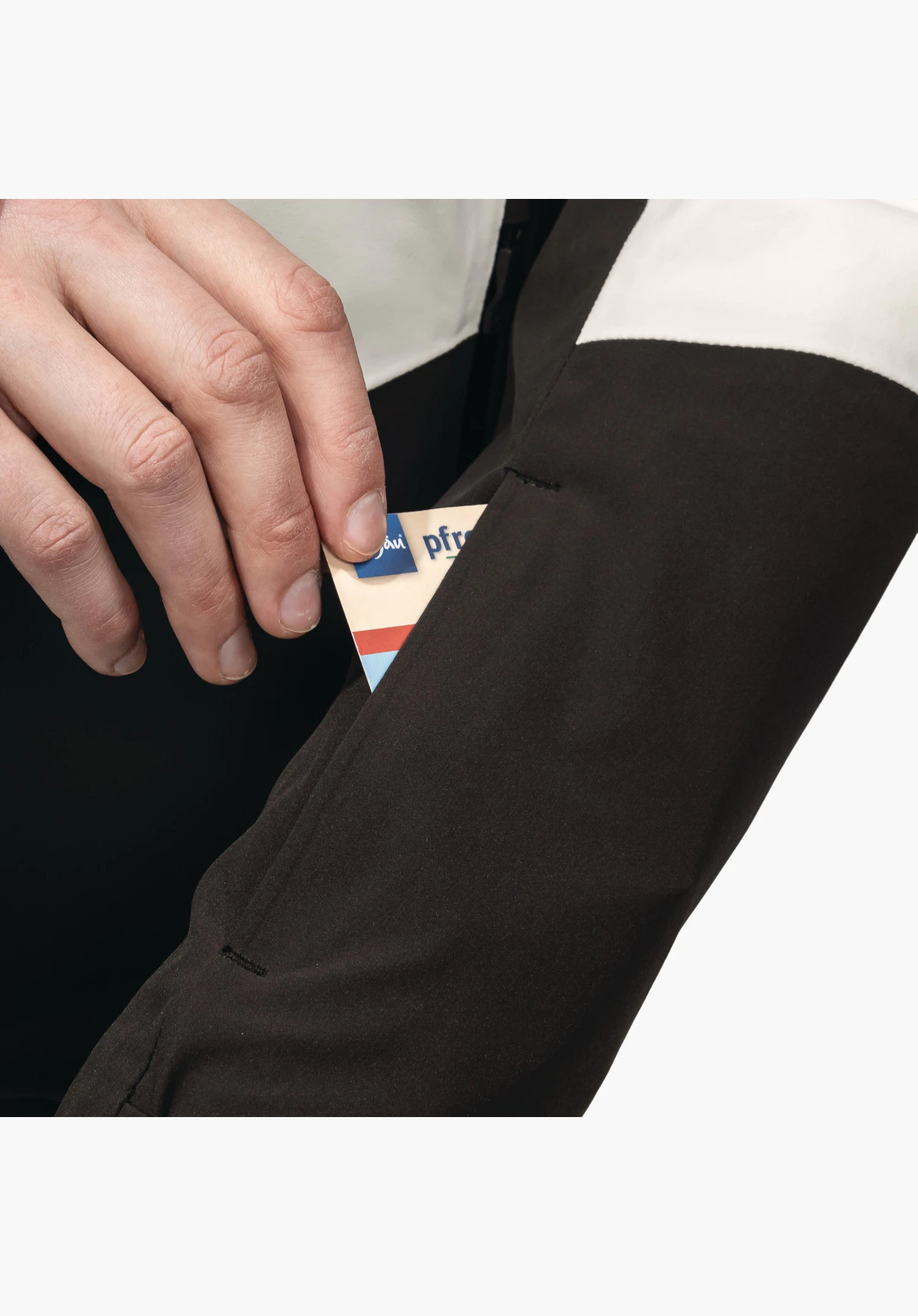 Jacket Style Safuna WMS toont de functionele skipaszak op de mouw waarin een hand een kaart schuift, uitgevoerd in zwart en natur materiaal, close-up tegen een witte achtergrond.