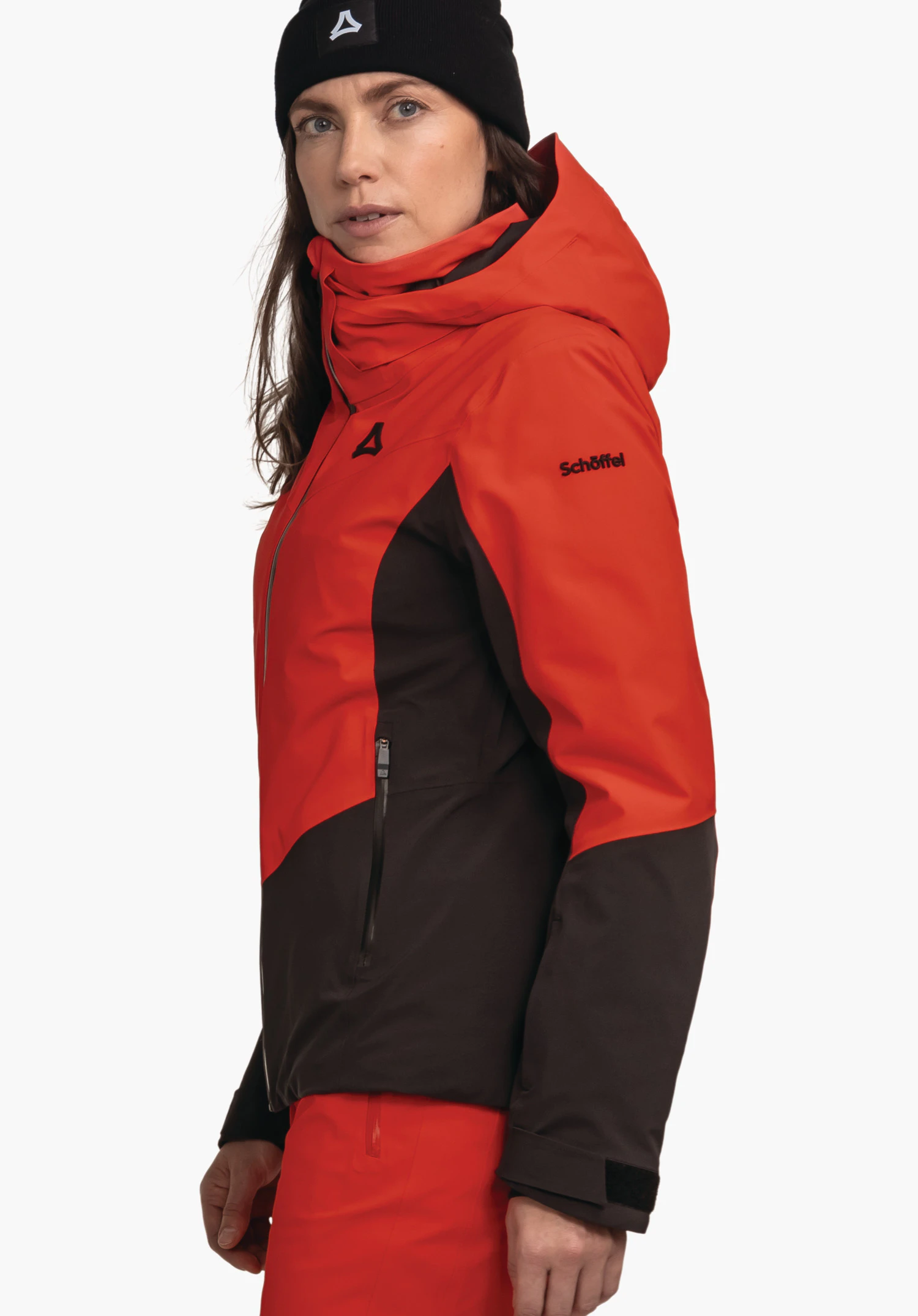 Jacket Style Safuna WMS rood-zwarte ski-jas met capuchon; vrouw draagt deze en kijkt zijwaarts; zwart beanie met Schöffel-logo; lichte neutrale achtergrond. Tekst: Schöffel.