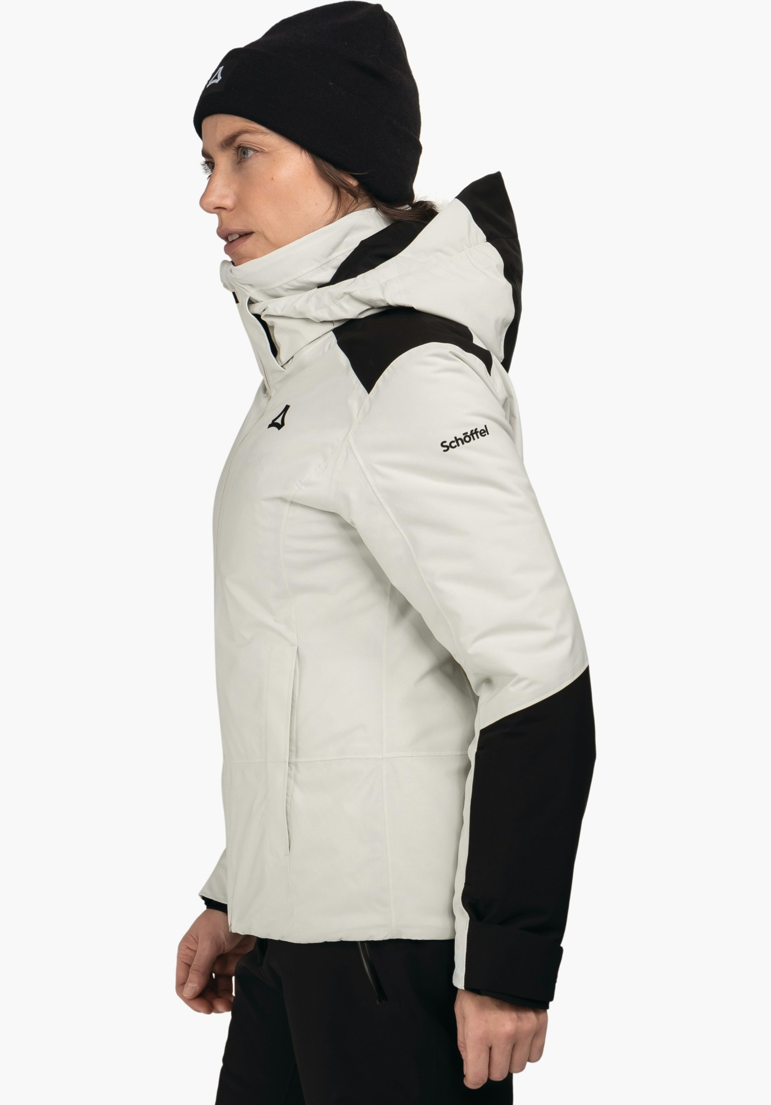Jacket Style Zandwel WMS femme portant une veste noire avec finitions blanc clair, souriante en profil, porte un bonnet noir et un pantalon vert foncé, debout devant fond blanc neutre.