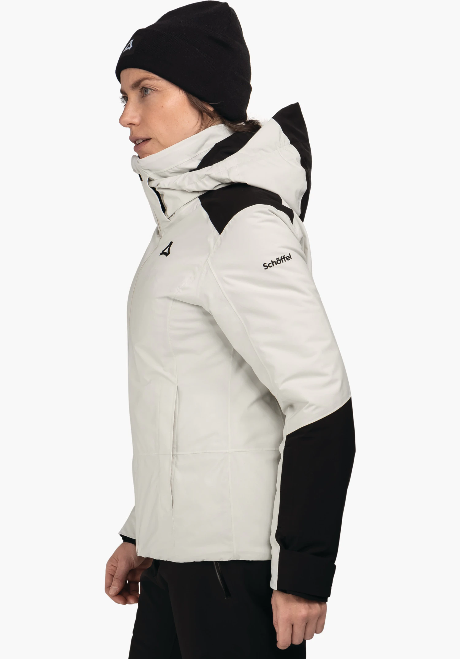Jacket Style Zandwel WMS femme portant une veste noire avec finitions blanc clair, souriante en profil, porte un bonnet noir et un pantalon vert foncé, debout devant fond blanc neutre.