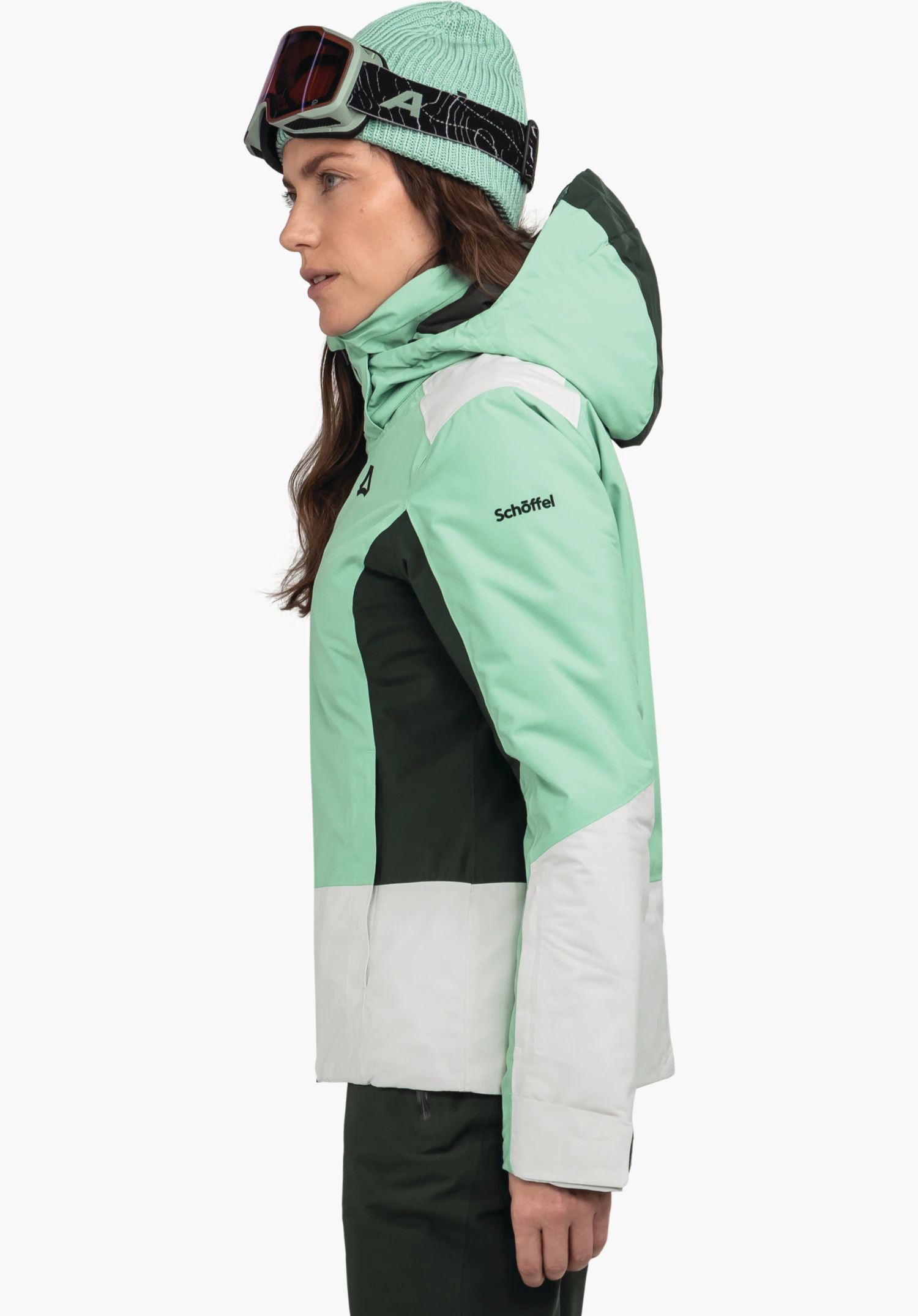 Jacket Style Zandwel WMS woman wearing a light green ski jacket with hood and dark green inserts, standing sideways and looking forward; wearing a light green beanie and ski goggles with black strap. Schöffel logo; text: "Schöffel".
