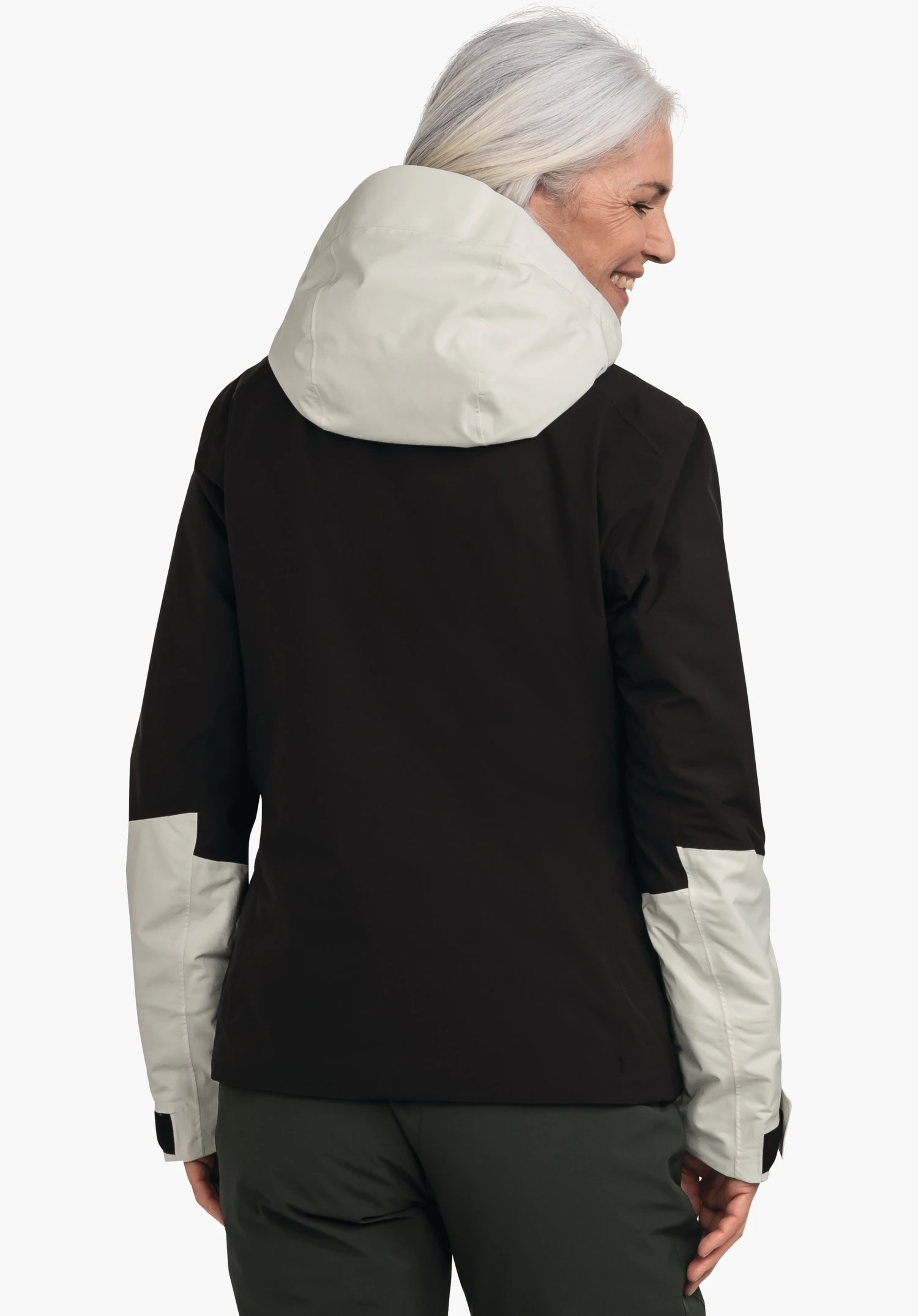 Jacket Style Trifex WMS zwarte jas met lichtgrijze capuchon en lichtgrijze mouwinzetten; vrouw met wit haar draait zich glimlachend om voor een witte achtergrond en draagt een donkergroene broek.